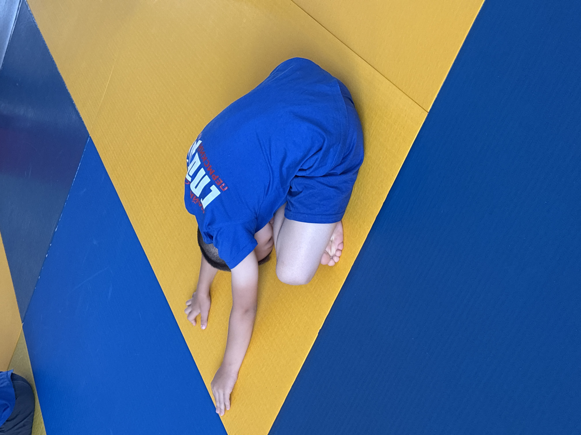 In a gymnasium, a young boy in a blue t-shirt and shorts is performing a seated forward bend exercise on a yellow mat. The mat is adjacent to a blue mat, creating a distinct visual contrast. The boy's arms are extended forward, and his head is lowered towards his knees. His bare feet are visible. The blue t-shirt has white text on the back that appears to be in Cyrillic script, possibly indicating a team or club name. The scene is likely taking place in Chaikovsky, Russia, a city known for its sports facilities.