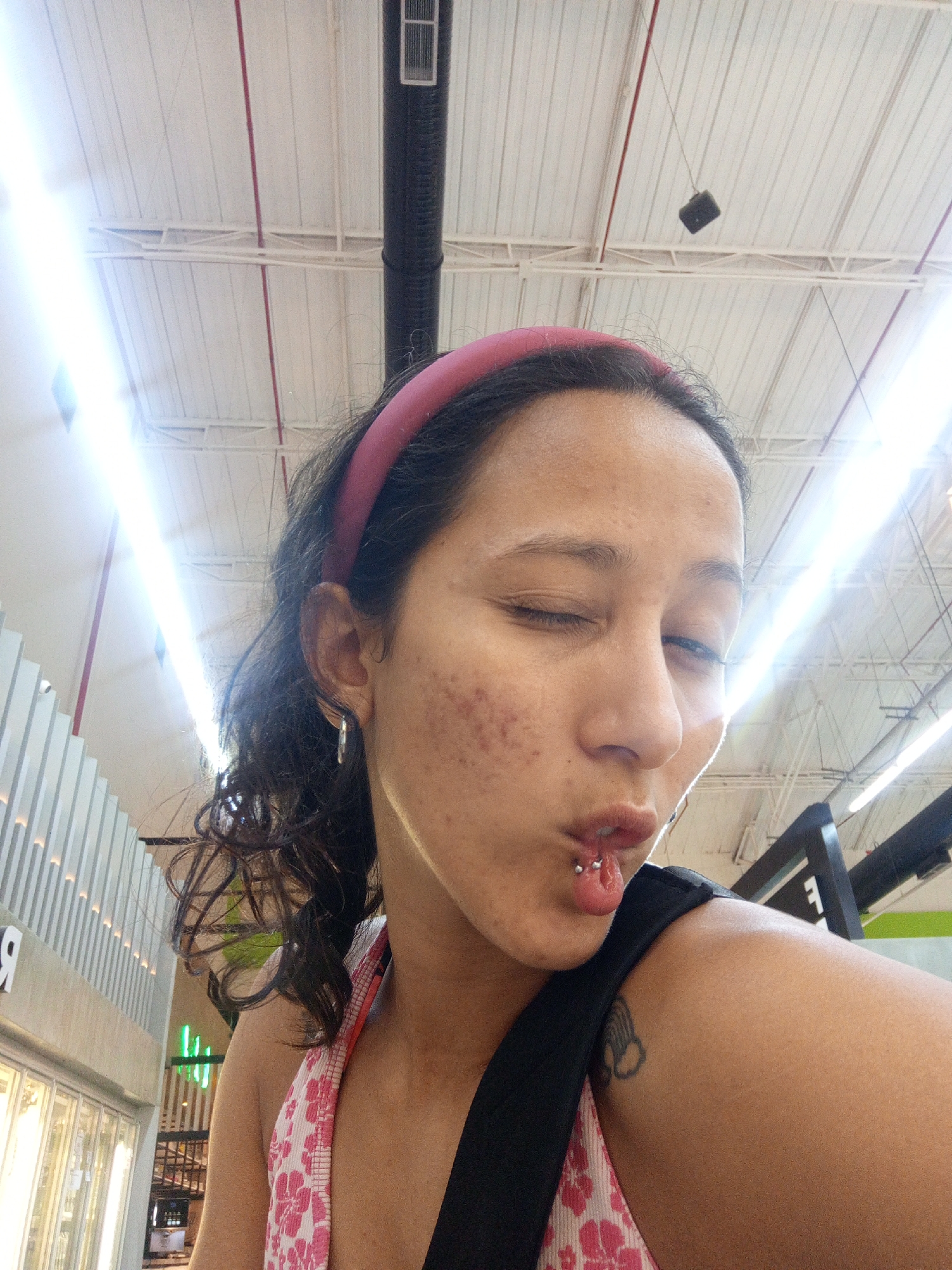 A woman with dark curly hair pulled back by a pink headband, her face partially turned to the side, is taking a selfie. She is winking with her right eye and sticking out her tongue, which is pierced with a horizontal barbell. She is wearing hoop earrings and a tank top with a floral print underneath a black strap. The background shows a commercial setting with a white corrugated metal ceiling featuring overhead lighting and visible ducts. The location is plausibly a commercial establishment in Porlamar, Venezuela.