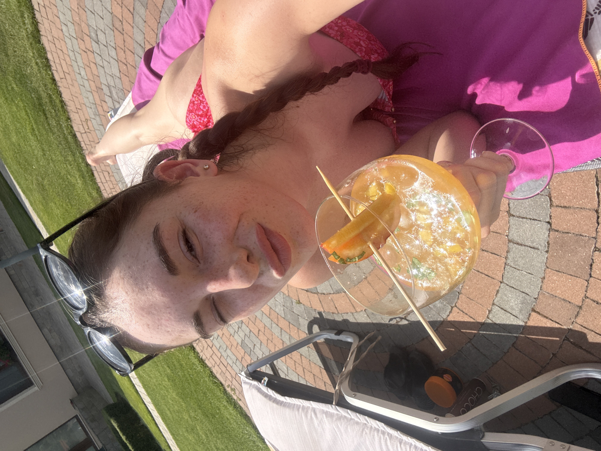 A woman is lying on a lounge chair outdoors on a sunny day. She is wearing a pink bikini and has her sunglasses on top of her head. She is holding a glass of an orange beverage with a slice of fruit and a mint sprig. The woman is looking at the camera with one eye closed and her lips pursed. The background shows a paved patio, a lawn, and a building. The city of Trud, Bulgaria, is not visible in the image, and there are no contextual clues to suggest this location.