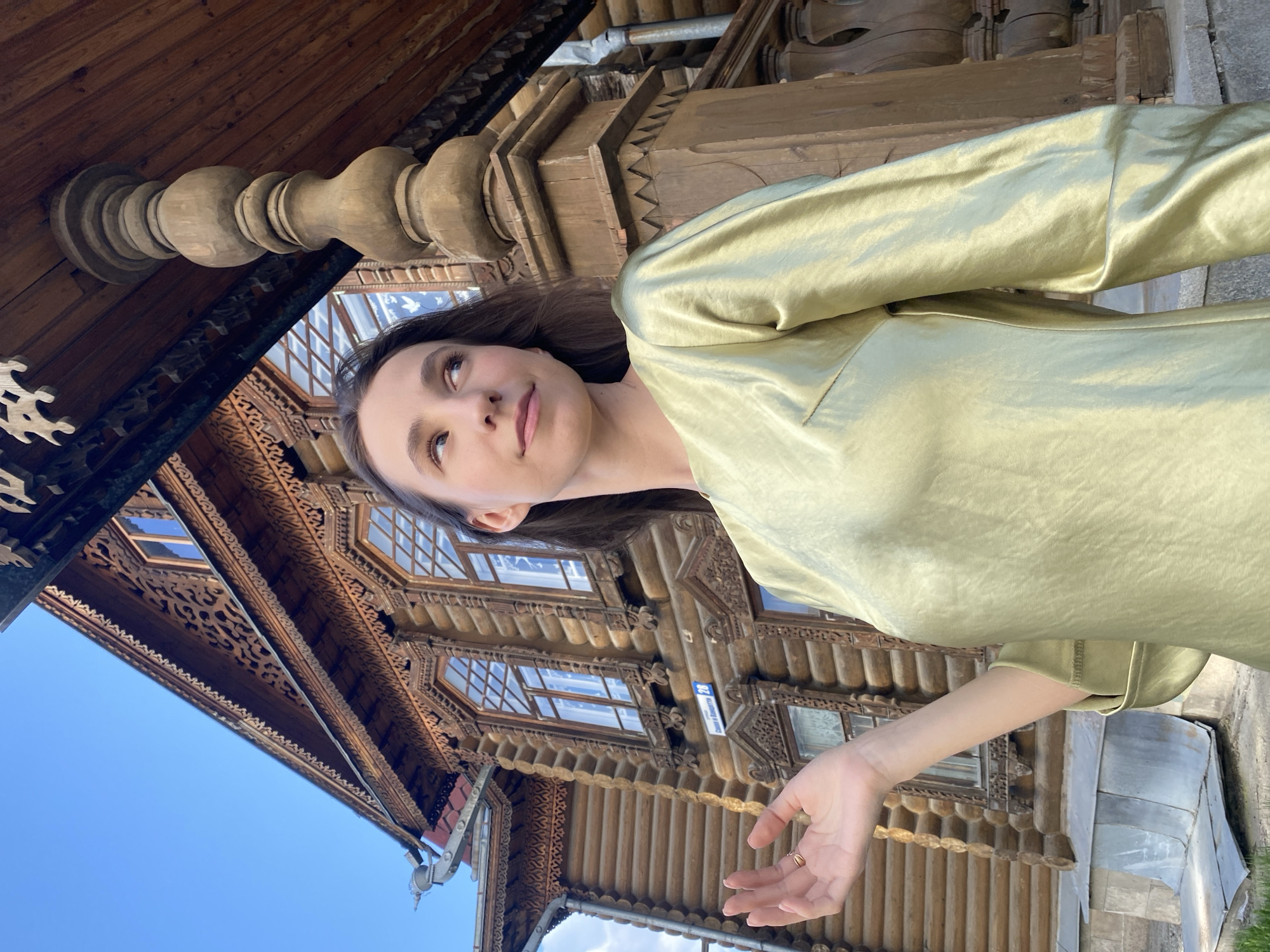 A woman is standing in front of the Sevastyanov House in Yekaterinburg, Russia. She is wearing a shiny, light green blouse or dress. Her dark hair is parted to the side and her face is turned slightly upwards and to her left. Behind her, the intricately carved wooden facade of the Sevastyanov House is visible. The sky is light blue and clear.