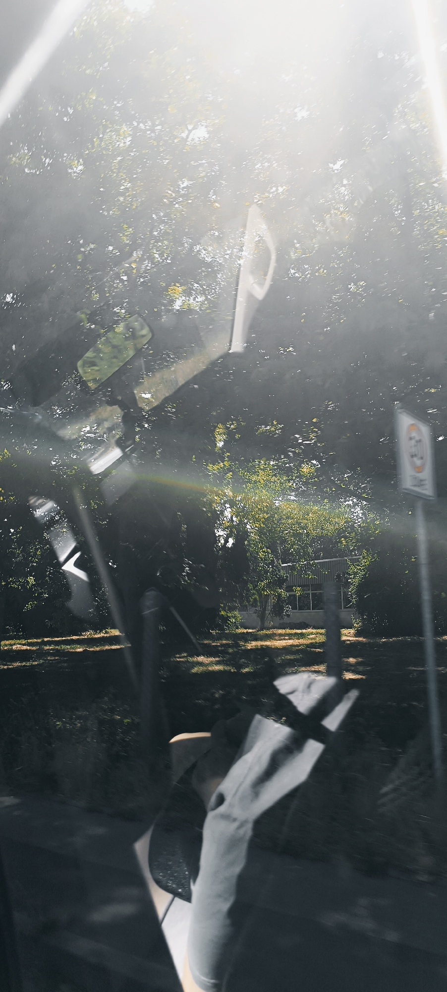 The image appears to be taken from inside a vehicle, looking out through the windshield or side window. The view is dominated by bright sunlight filtering through a dense canopy of green trees, creating lens flares and a hazy, overexposed effect in the upper portion of the frame.
In the foreground, several reflective surfaces, likely parts of the vehicle's interior such as the rearview mirror, are visible and create distorted reflections of the outside environment. There are also some abstract, light-colored shapes that may be parts of the car's dashboard or other interior components.
Further out, through the greenery, a small, light-colored building with several windows is visible. To the right of the building, a traffic sign is partially visible. It is a circular sign with a red border and a white background, featuring a black silhouette of a vehicle. Inside the circle, there is a red bar across the top. Below the circle, there appears to be text, possibly indicating a speed limit.
The ground visible beneath the trees appears to be a mix of dry grass and dirt. The overall impression is a sunny day, possibly in a semi-rural or suburban area with vegetation, and the presence of the traffic sign suggests a road or public area. The lens flares and reflections indicate that the sun is positioned behind the viewer, or at an angle that causes significant glare.
It is not possible to definitively identify the location as Winterbach, Germany, based solely on the visual information in this image.
