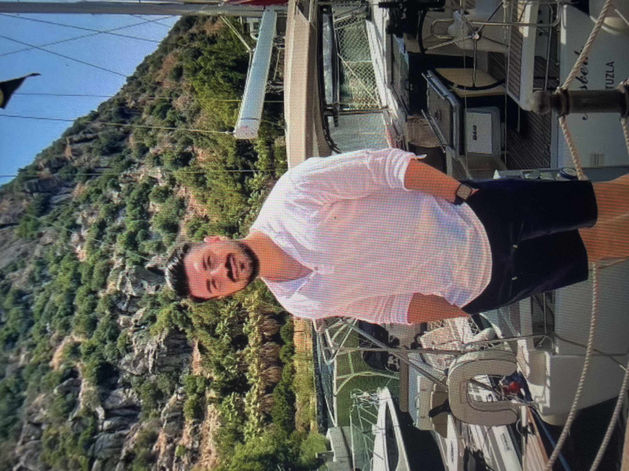 The image depicts a male subject standing on a dock in a marina setting. The man is positioned centrally, facing the viewer, wearing a white long-sleeved shirt with rolled-up sleeves and dark shorts. He has dark hair and a beard, and his hands are in his pockets.
In the midground, several sailboats and yachts are moored. Partial views of their sterns, cabins, and rigging are visible. On the stern of a white yacht in the lower right, the partial text "...sbert." and "TUZLA" is discernible. A white horseshoe life buoy is attached to the stern of a boat on the left. A thick rope runs horizontally across the lower foreground, suggesting a mooring line or part of the dock's infrastructure. A dark wooden bollard or post is visible in the bottom right corner.
The background features a steep, verdant hillside or mountain covered with dense green vegetation, including trees and rocky outcrops. The sky above the mountain is clear and blue. The overall environment suggests a coastal or large lake marina with mountainous surroundings.