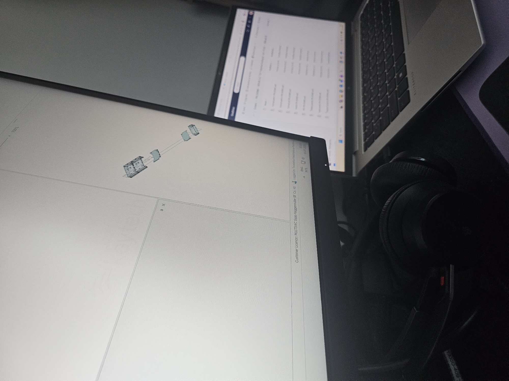 An overhead, slightly angled shot captures a modern workspace featuring a large computer monitor displaying a 3D model and a laptop open to a file explorer window. The monitor, positioned on the left, shows a technical schematic with the text "keytech" visible in the upper portion. The lower edge of the monitor displays "Customer Licence: MULTIVAC Sepp Haggenmüller SE Co. KG" and "Logged in: Tsvetelina Pokharanova Pașliev." A pair of black over-ear headphones rests on a dark surface in the foreground, to the left of the laptop. The laptop, situated on the right, is a silver HP EliteBook, displaying a directory of PDF files. The environment appears to be indoors, likely an office or home office, with artificial lighting casting subtle shadows. The scene is devoid of any individuals and suggests a work-in-progress or a moment of paused productivity. No identifiable landmarks or specific features indicate the city of Sofia, Bulgaria.