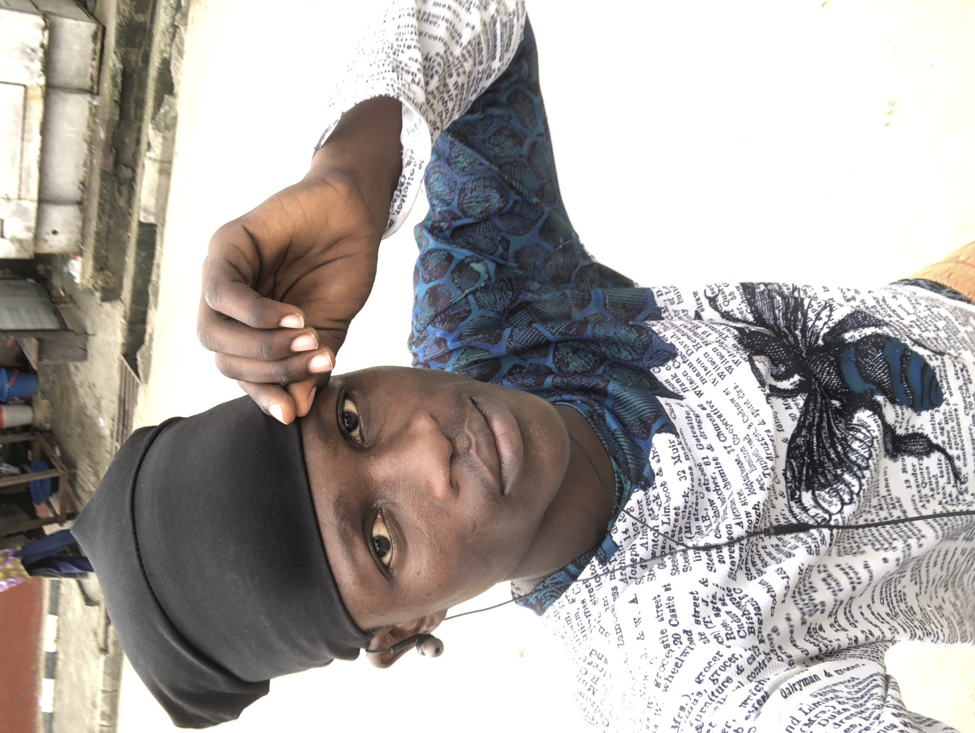 A medium shot captures a young Black male, possibly in his late teens or early twenties, posing for a selfie outdoors in what appears to be a street scene in Ado, Nigeria. He is wearing a black skullcap and a patterned shirt with a white background featuring text and an illustration of a large insect. The shirt's design includes a blue and green section with a scale-like pattern. He has a wired earbud in his left ear and is touching his forehead with his right hand, his fingers slightly curved. The background is blurred, but suggests an urban environment with a street, a sidewalk with a curb, and what looks like a building structure behind him. The overall lighting indicates it is daytime.