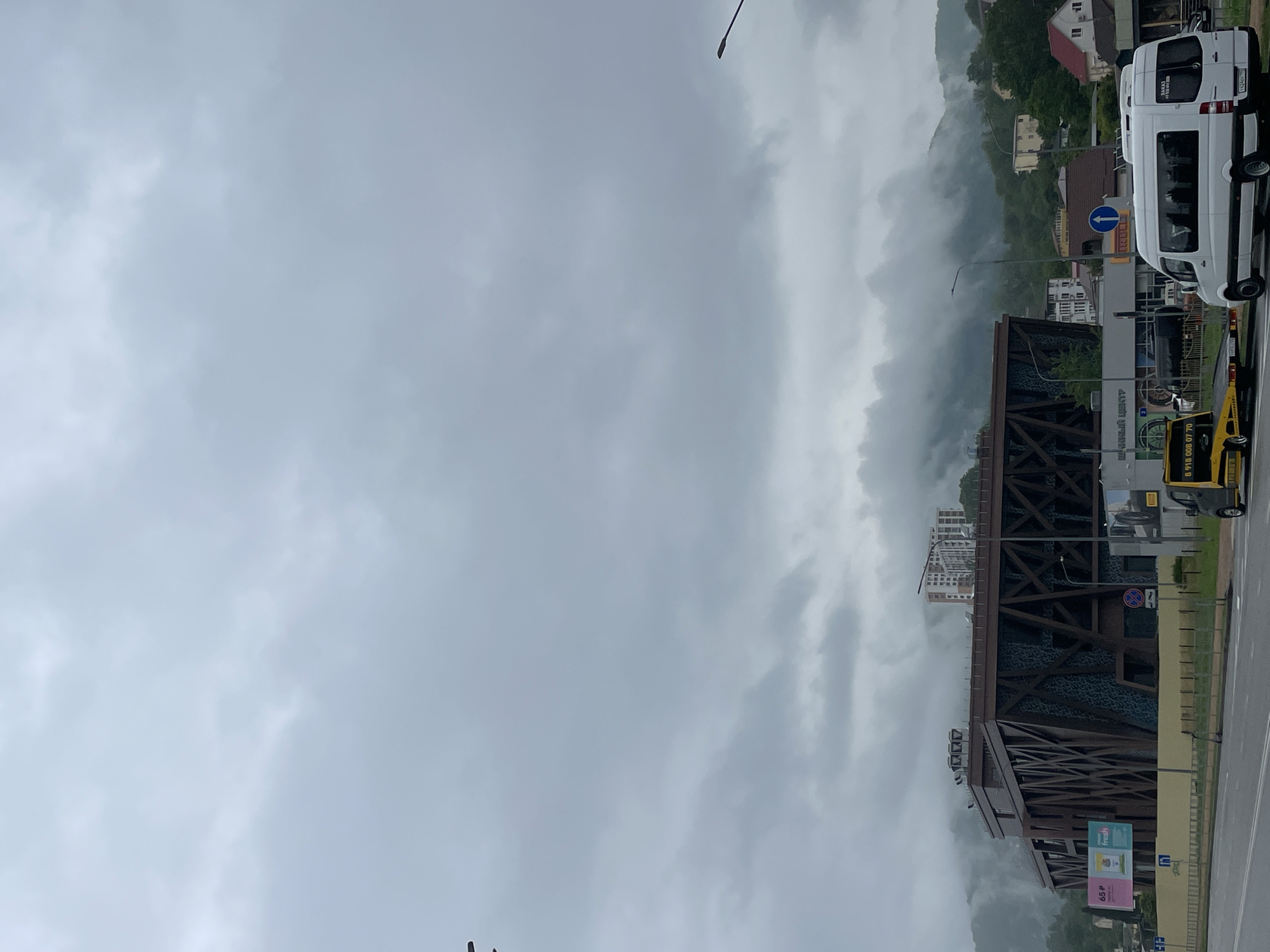 The image captures a cloudy day in Sochi, Russia, featuring a building with a prominent structural framework and textured cladding. A white van and a yellow taxi are parked along a roadside adjacent to the building. The backdrop is a hillside partially obscured by mist, with residential buildings visible. Traffic signs are present near the building and vehicles.