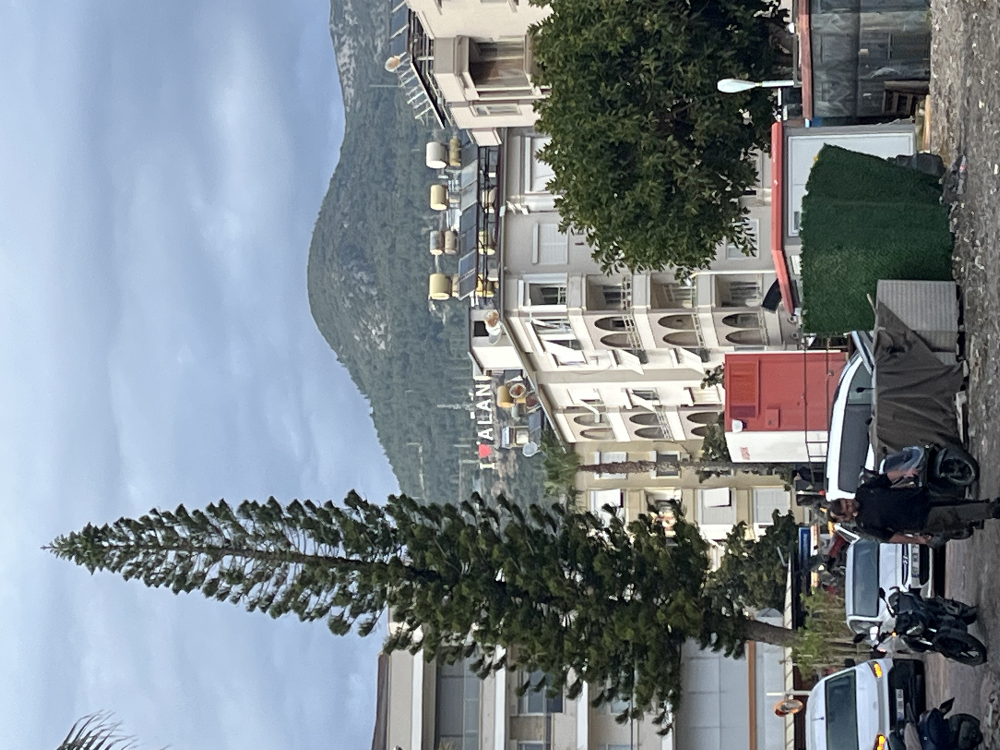 The image shows a street in Alanya, Turkey. In the background is a mountain range. A tall, slender pine tree is in the foreground on the left. Several multi-story buildings line the street, with an "I LOVE ALANYA" sign visible on one. Vehicles, including cars, vans, and scooters, are parked along the street. A person is standing near the parked scooters. The sky is overcast.
