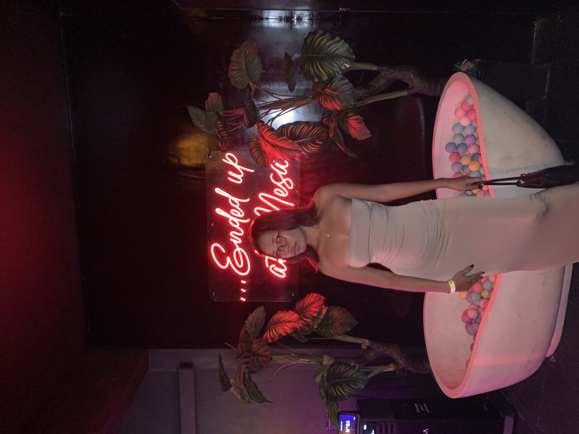 The image shows a person standing in a dark indoor location, likely a club or lounge, in North Kuta, Indonesia. The person is wearing a strapless dress. Behind them is a wall decorated with artificial plants and a neon sign that reads "...ended up at Mesa". To the right, there's an oversized egg-shaped structure filled with colorful balls. The person appears to be interacting with the egg-shaped structure. The environment is dimly lit with neon lighting effects.