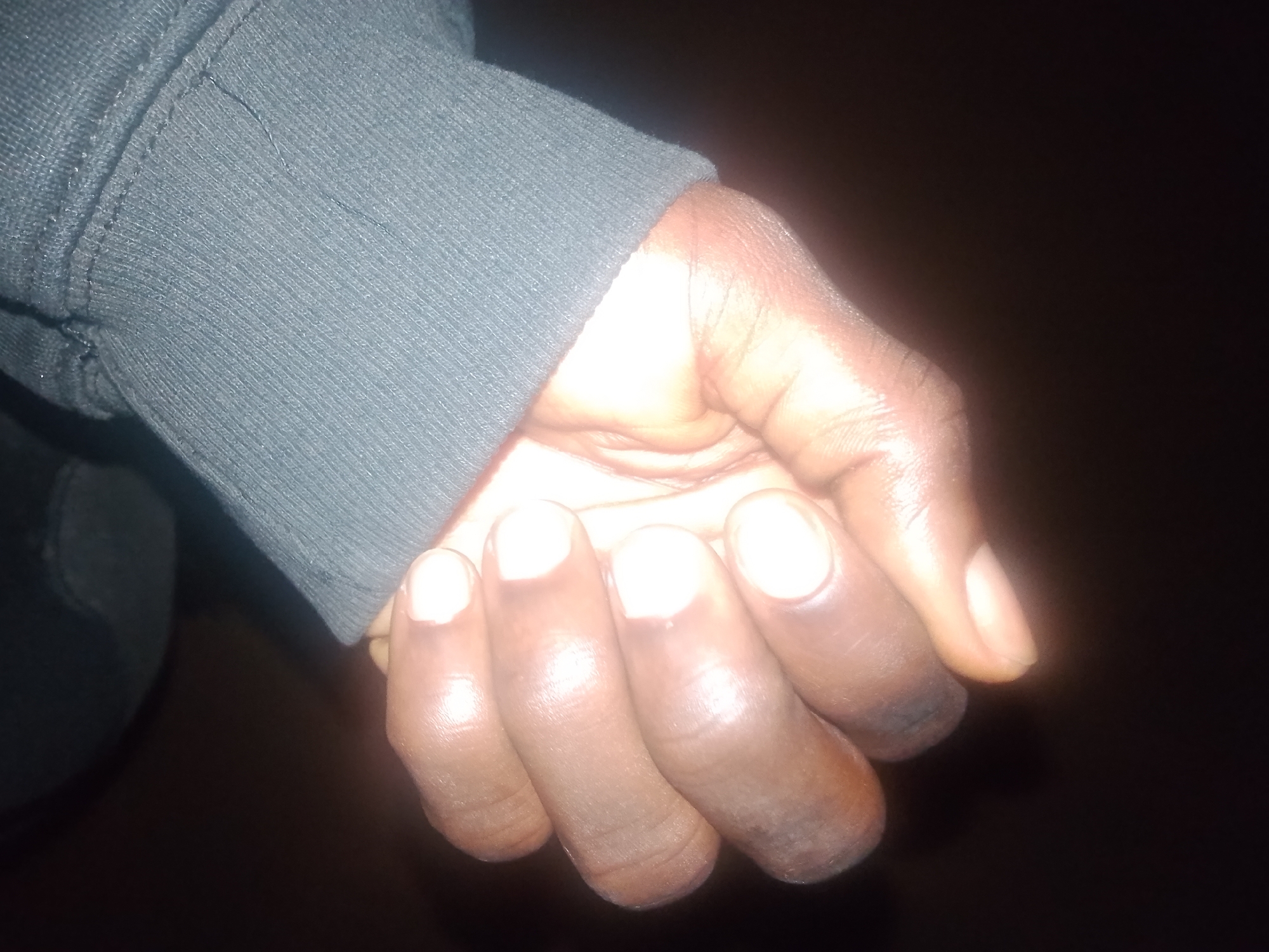 The image depicts a close-up view of a person's hand, partially covered by the sleeve of a dark-colored garment, likely a sweatshirt or jacket. The hand is positioned against a dark, out-of-focus background. The lighting suggests it was taken indoors or in a dimly lit environment, highlighting the texture of the skin and the garment. The hand appears to be held in a relaxed, slightly curled position, with the fingers visible. The skin tone is dark, and there are visible wrinkles and creases on the fingers, indicative of natural skin texture. The garment's sleeve has a ribbed texture, typical of cuffs on casual wear. The overall scene is simple, focusing solely on the hand and a portion of the clothing. There is no discernible information in the image that would place the location in Jalingo, Nigeria, or any other specific city.