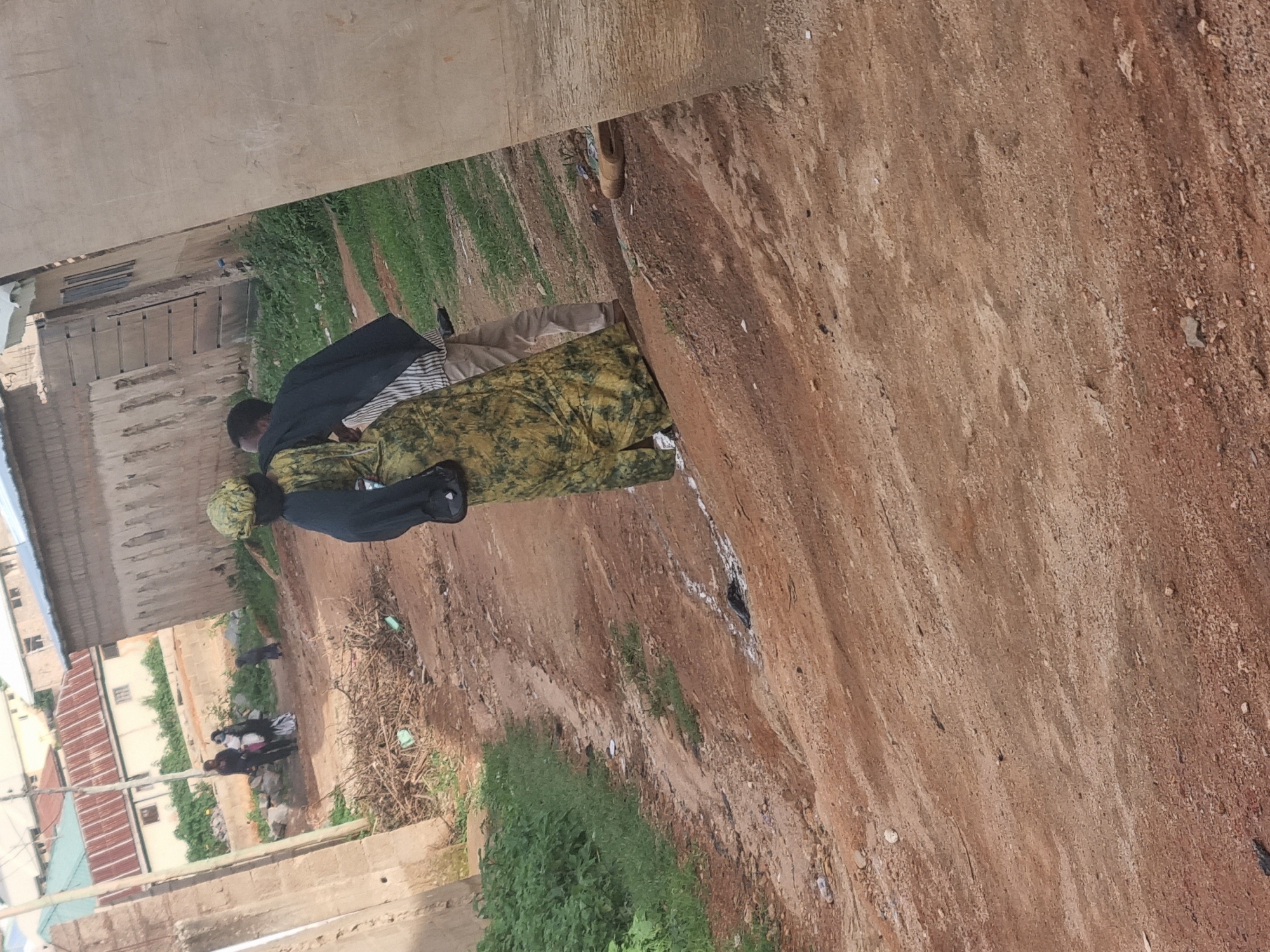In Jos, Nigeria, a dirt road winds through an urban area. Two individuals, a woman in a green and yellow patterned dress and a man in a black jacket and striped pants, walk away from the camera. They appear to be in a casual setting, perhaps strolling through their neighborhood. Buildings, some appearing unfinished, line the road, suggesting ongoing development or a mix of older and newer structures. The landscape is predominantly dry and earthy, with patches of green vegetation suggesting a semi-arid climate. The scene captures a slice of everyday life in this part of Jos, showcasing the local architecture and the natural environment.