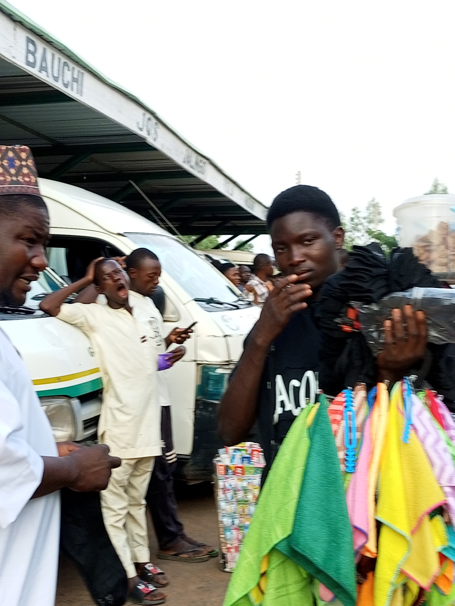 In Gombe, Nigeria, at what appears to be a motor park or roadside market, a group of men are gathered around a white commercial bus. A vendor, wearing a black t-shirt with the partial word "ACO" visible, stands in the foreground, holding a bundle of black plastic-wrapped items and displaying a rack of colorful folded cloths, possibly towels or handkerchiefs. To his left, a young man in a cream-colored tunic is leaning against the bus, yawning widely with his hand on his head, and holding a smartphone and a small purple cup. Another man in a dark shirt stands behind him. Further left, a man wearing a patterned cap and a white garment is partially visible, looking towards the scene. In the background, the word "BAUCHI" is painted on the white roof of an overhang, suggesting a connection to that city or region. The environment is bright and appears to be outdoors, with trees visible in the distance and a clear, overcast sky. The overall scene depicts a busy commercial transaction or a moment of rest and interaction at a transportation hub.