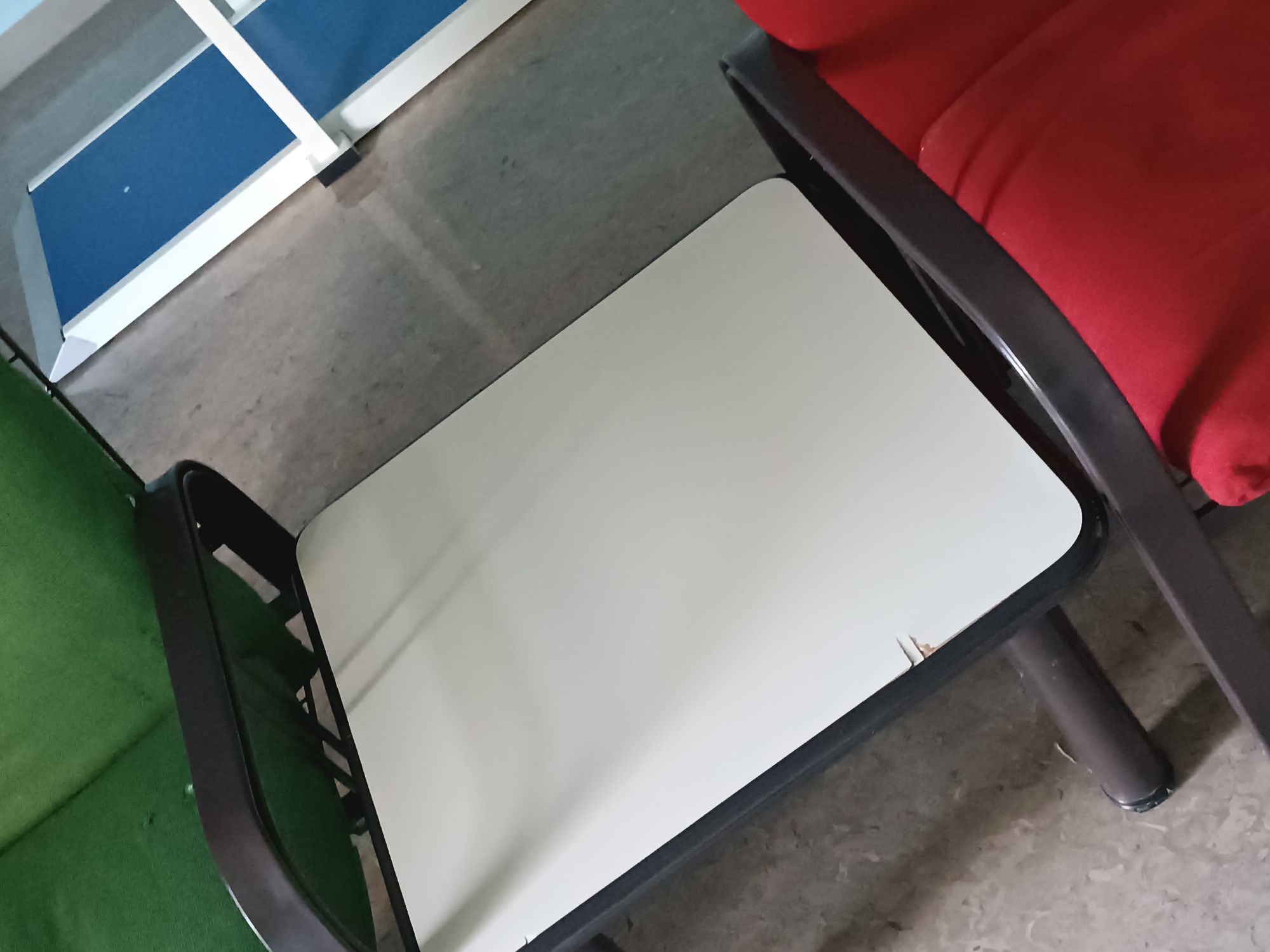 The image shows a close-up of a white tabletop with a black frame, resting on dark brown legs. The tabletop is slightly tilted and has some minor damage on its surface. To the left of the tabletop, a green upholstered chair is partially visible. To the right, a portion of a red upholstered chair is visible. In the background, a white shelf unit with blue panels can be seen. The flooring appears to be concrete. The scene is indoors, possibly in a waiting area or a classroom. There are no people present in the image. The lighting suggests an indoor environment, potentially with overhead lighting.