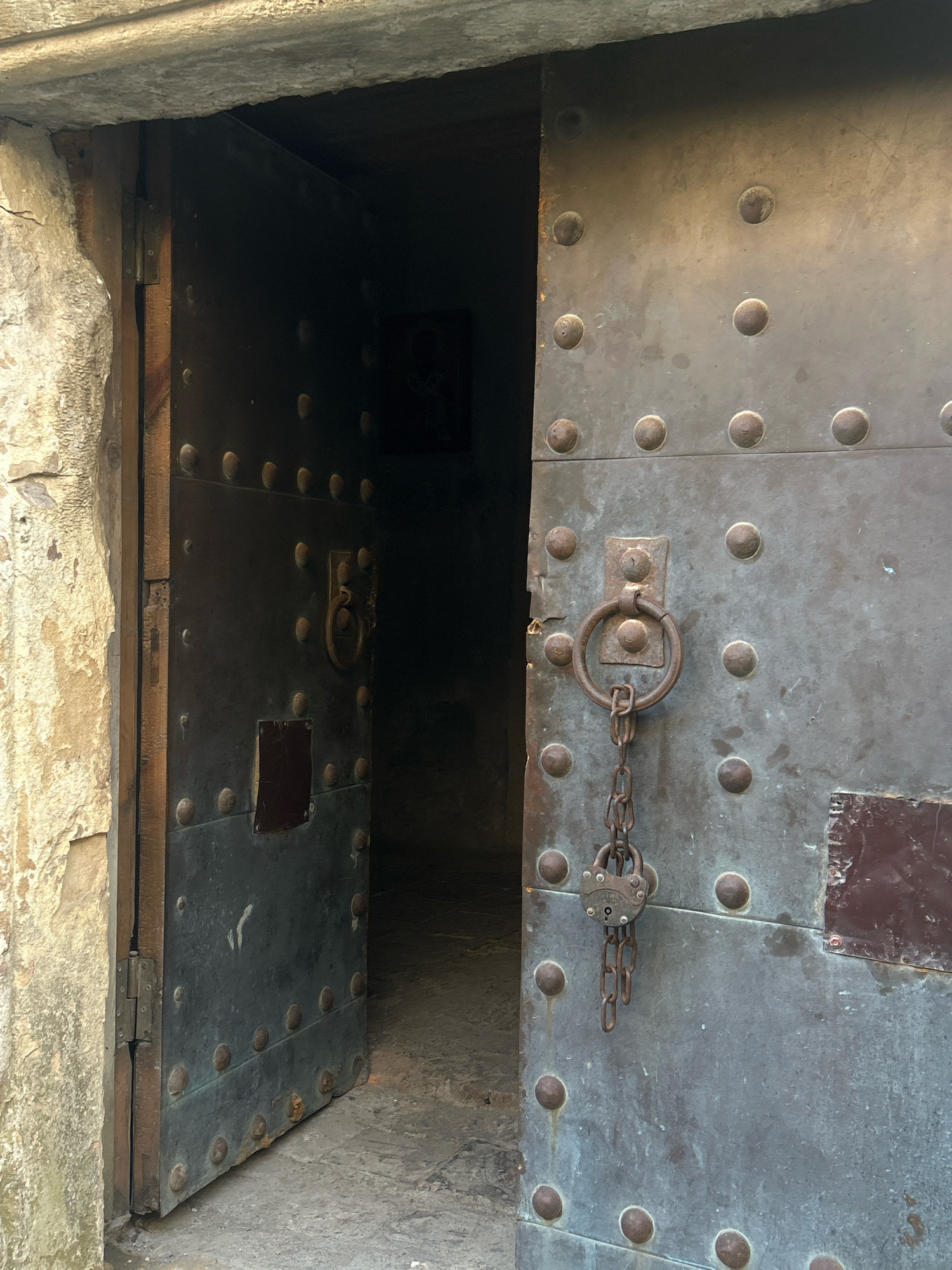 The image shows a partially opened, heavy, metal door, characteristic of old fortifications, set within a stone entryway. The door is reinforced with numerous round rivets. A circular metal handle is attached to the door, with a rusty chain leading to a padlock that is secured. The door is hinged to a wooden frame, which is set into a rough, stone wall. The interior visible through the open doorway appears to be dimly lit and suggests a passage or room beyond. The scene is likely located in Shalikiantkari, Georgia, a city known for its historical structures and fortresses. The image captures a moment of stillness, with no people present, emphasizing the age and sturdiness of the entrance.