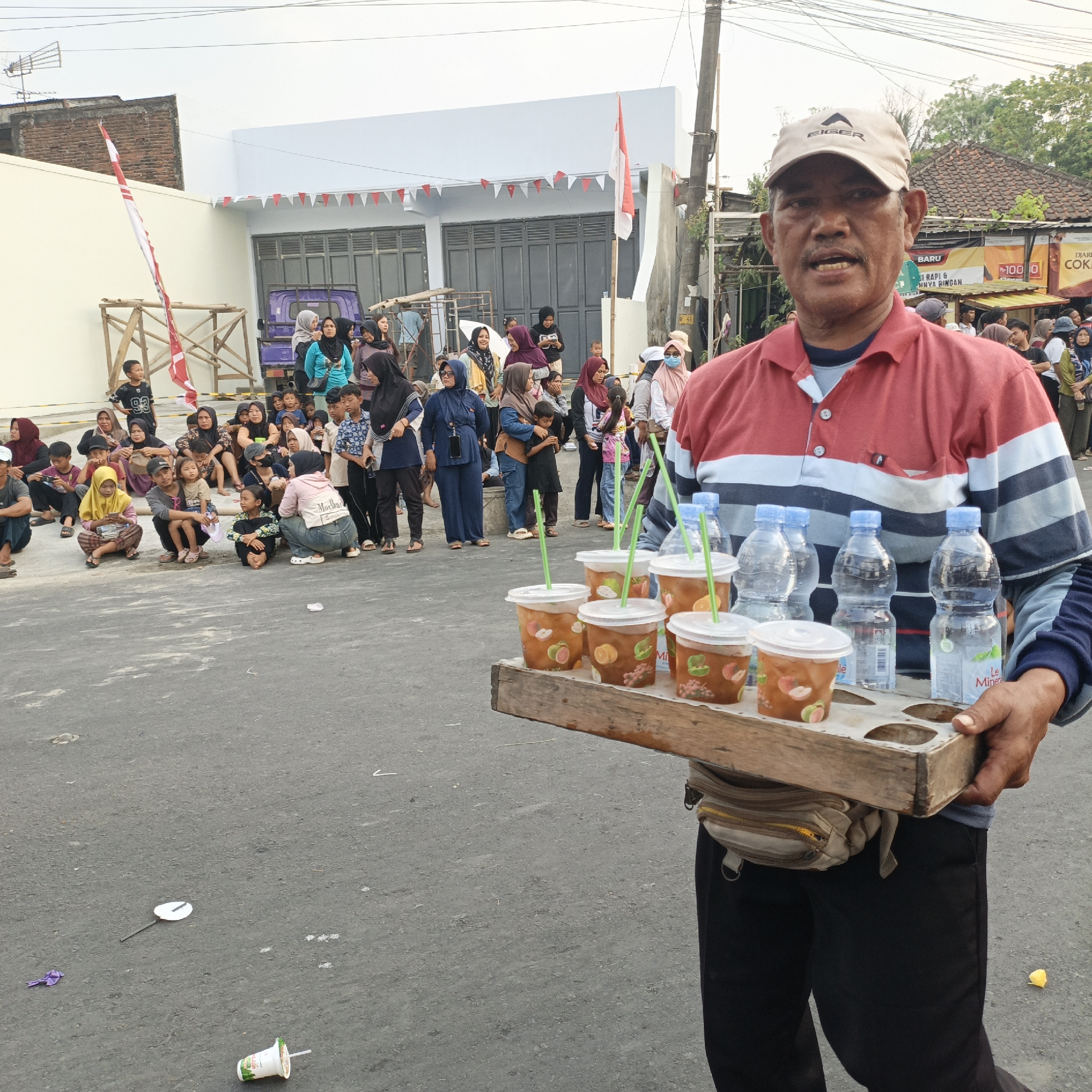 A man in a red and blue striped polo shirt and a beige cap holds a wooden tray with six plastic cups of iced drinks and four plastic water bottles. The cups have green straws and images of fruit. The man is standing on a gray asphalt road, with a crowd of people, many of whom are wearing headscarves, sitting and standing behind him. In the background, there is a white building decorated with red and white triangular flags, a flagpole with an Indonesian flag, a purple truck, and other buildings and trees. The scene appears to be a street gathering or event. The context of Randudongkal, Indonesia, suggests a local community event or market setting. There is some litter on the ground, including a plastic glove, a small white fan on a stick, and a discarded cup with a straw.