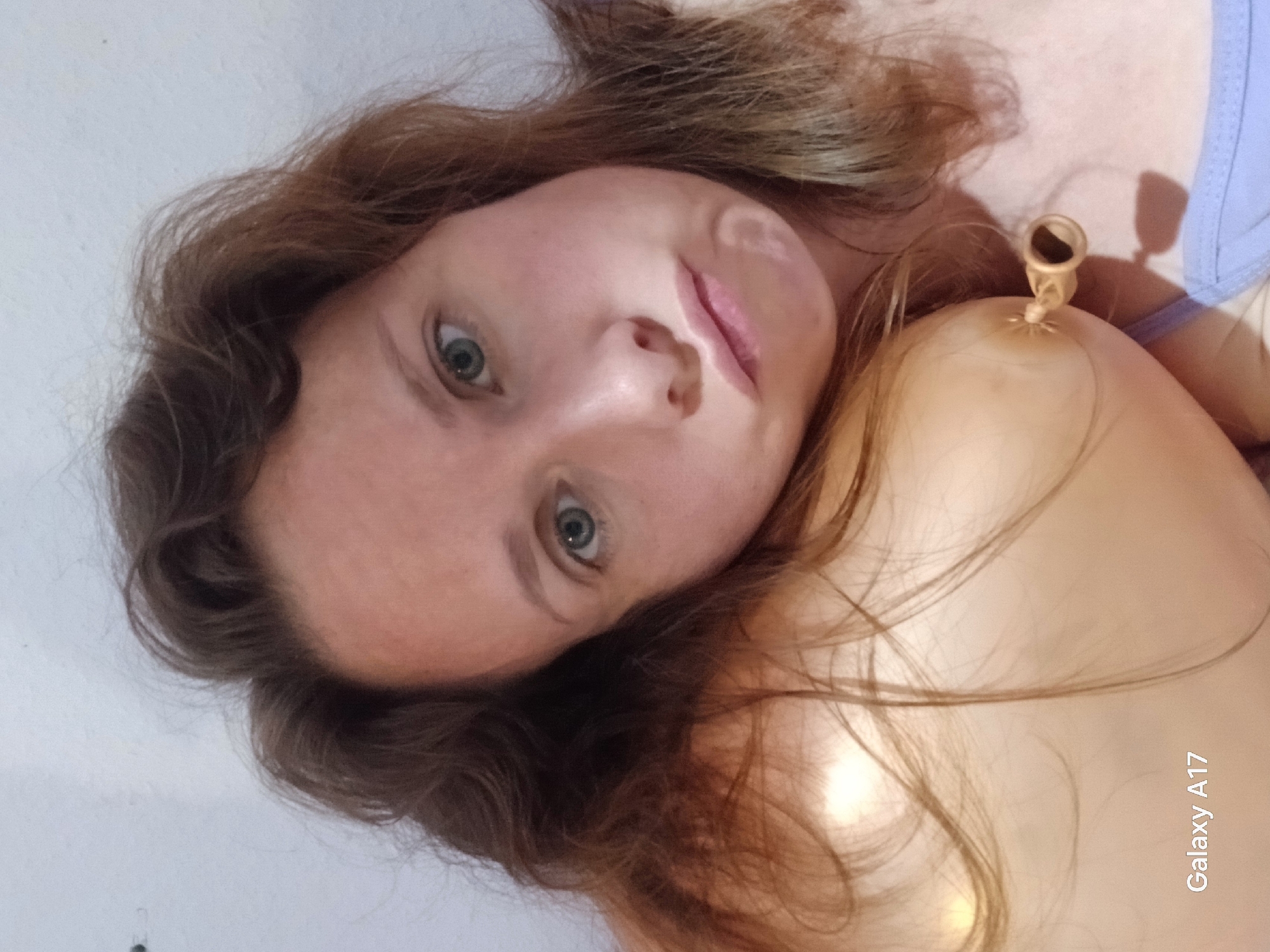 The image is a close-up portrait of a light-skinned female, oriented 90 degrees clockwise, with her head positioned on the left and her upper torso extending towards the right. She has long, light brown or auburn hair, appearing somewhat tousled around her face and shoulders. Her eyes are a striking shade of hazel or green, wide open and gazing directly into the camera with a neutral to slightly curious expression. Her lips are slightly parted.
She appears to be lying down, with her head resting against a plain, light-colored surface that forms the background of the image, possibly a wall or bedding. Near her right shoulder/collarbone area, a small, light-colored, elongated object resembling a deflated balloon or a specific type of medical device (like a small cupping cup without suction) is visible. A sliver of a light blue garment, possibly a bra strap or top, is visible on the far right edge of the frame.
The lighting is soft and diffused, characteristic of an indoor setting. No specific time of day or outdoor weather conditions can be determined from the image. There are no explicit location cues within the image to suggest Yekaterinburg, Russia. The emotion conveyed is one of calm engagement.
The text "Galaxy A17" is visibly watermarked in white letters, oriented vertically along the right side of the image, indicating the mobile phone model used to capture the photograph. The overall activity is a direct gaze at the camera in a relaxed, intimate setting.