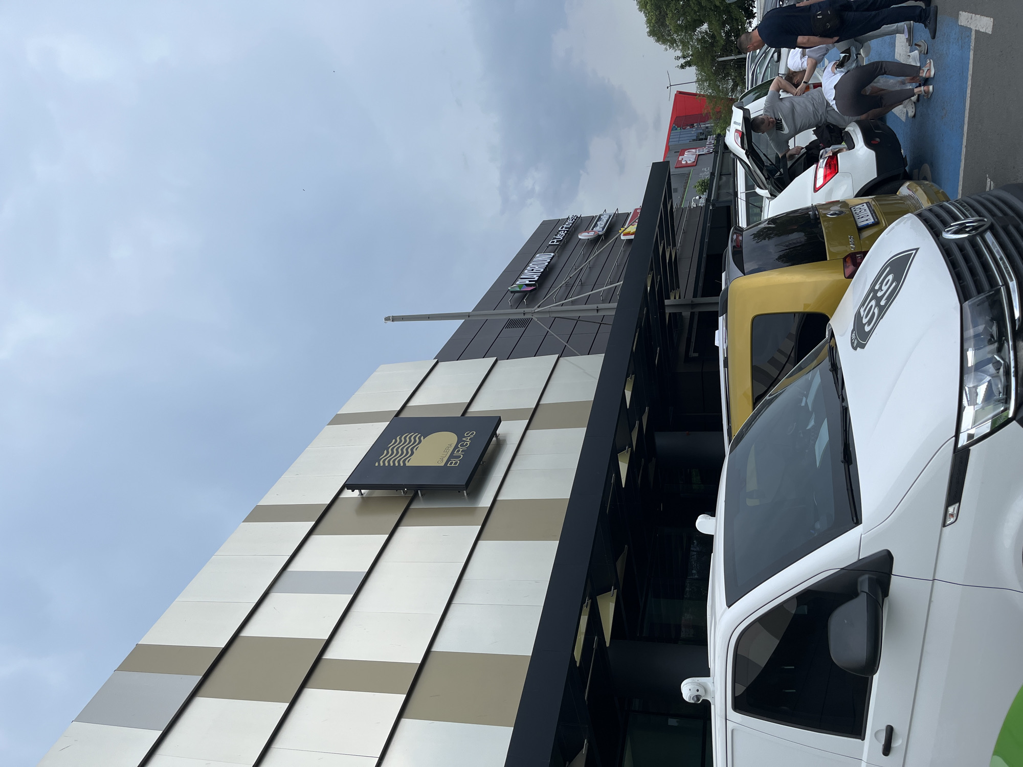 A ground-level, angled shot focuses on the "Galleria Burgas" shopping center in Burgas, Bulgaria. The building's exterior features alternating horizontal stripes of silver and gold panels. A dark sign displaying the name "GALLERIA BURGAS" is attached to the facade. Above, a sign displays "PLAYGROUND Pulse Fitness". A white van is parked in the foreground. To the right and rear of the van, a yellow car and a white car with an open hatchback are visible. Several people are interacting with the open hatchback. The sky above is overcast. The lighting is natural.