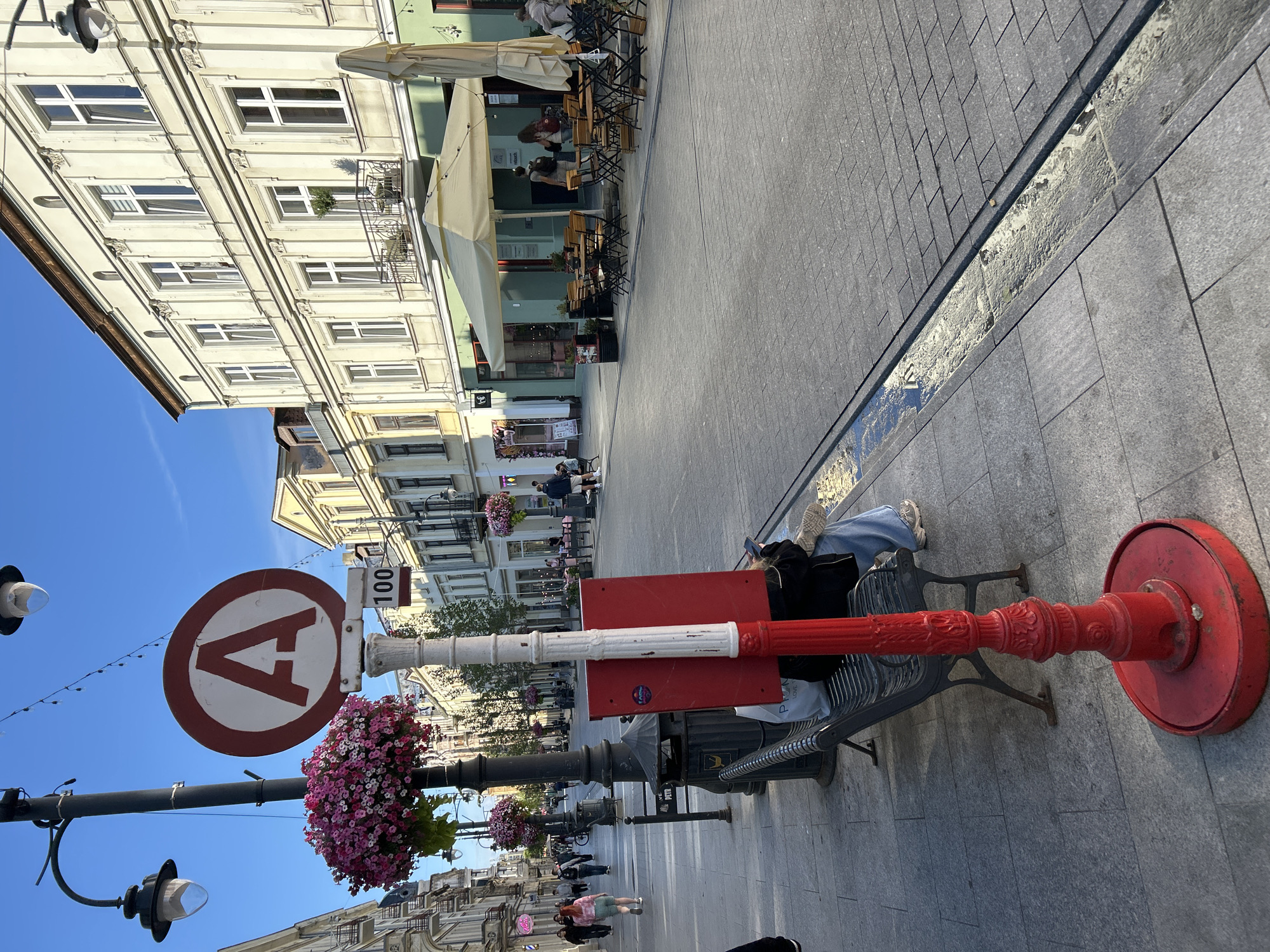 The image depicts a street scene in Łódź, Poland, likely during the daytime, given the bright sunlight and shadows. The foreground features a red-and-white striped lamppost base with a red sign attached to it. To the left of the lamppost is a metal bench where a person is seated, facing away from the camera, looking at their phone. The person is wearing blue jeans and light-colored sneakers. A black shopping bag with white lettering is visible on the bench.
The street itself is paved with gray cobblestones. A narrow drainage channel runs along the edge of the sidewalk. In the background, a street lined with buildings is visible. These buildings have architectural details typical of European cities, with multiple stories, large windows, and ornate facades. Some buildings house businesses, with awnings and outdoor seating for cafes or restaurants.
There are several lampposts along the street, one of which has a hanging basket overflowing with pink and white flowers. People are seen walking along the sidewalk and sitting at outdoor tables. A circular red-and-white sign with a large red "A" in the center is mounted on a pole further down the street, accompanied by a smaller rectangular sign displaying "100". The sky is clear and blue. The overall atmosphere is that of a lively urban environment.