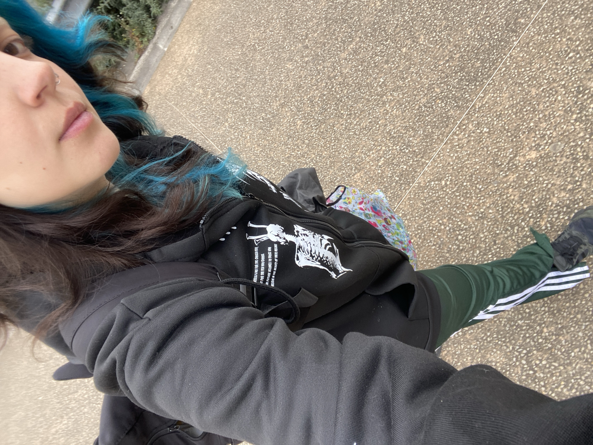 The image presents a partial view of a person outdoors in Gijón, Spain, captured from a high-angle, downward perspective, likely a selfie. The subject has dark hair with vibrant blue highlights and a nose piercing. They are dressed in a black jacket or hoodie, and a black fanny pack or crossbody bag is worn across their chest. The bag displays a white graphic depicting a skeletal figure and accompanying text. Partially visible to the right of the bag is a colorful item with a floral pattern, possibly a bag or scarf. The person is also wearing dark green athletic pants with distinctive white stripes along the sides, and black athletic shoes.
The setting is a paved outdoor area, characterized by a light brown and grey speckled surface, suggesting a walkway or urban path. Green shrubbery or foliage is visible in the background on the left. The natural light indicates it is daytime, with overcast conditions suggested by the lack of harsh shadows. The person's expression appears neutral.
Text visible on the fanny pack, as identified by OCR, reads: "ACCESS'S NOT FOR THE CHOSEN FEV, IT FOR THE FEW WRO CHOOSE THE FUTURE BELONGS TO THOSE VRO PETERE IN THE BEAUTY OF THEIR DREAMS".