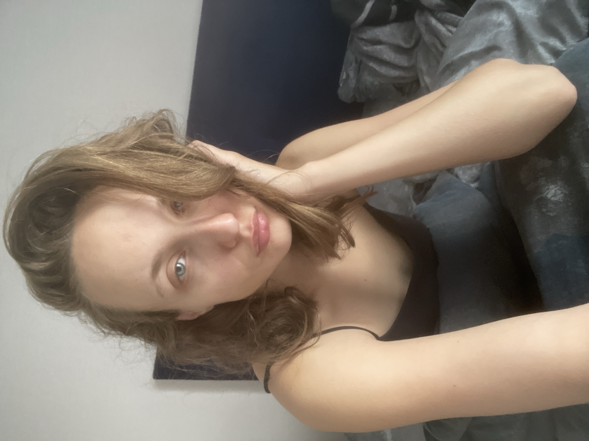 The media file is a close-up, portrait-style selfie of a young woman with light brown hair and blue eyes. She is lying down on a bed with gray bedding and a dark blue headboard visible in the background. Her hair is slightly messy and framing her face. She is wearing a black camisole with thin straps. One hand is raised and touching her hair near her temple, while the other arm is resting on the bed. The lighting suggests it could be daytime, possibly indoors with natural light. The woman's expression is neutral, with her eyes open and looking towards the camera. Her lips are slightly parted and appear to have a light pink tint. The overall setting is intimate and casual, suggesting a personal moment. No text is visible in the image. The location context provided is Подшивалово, Russia.