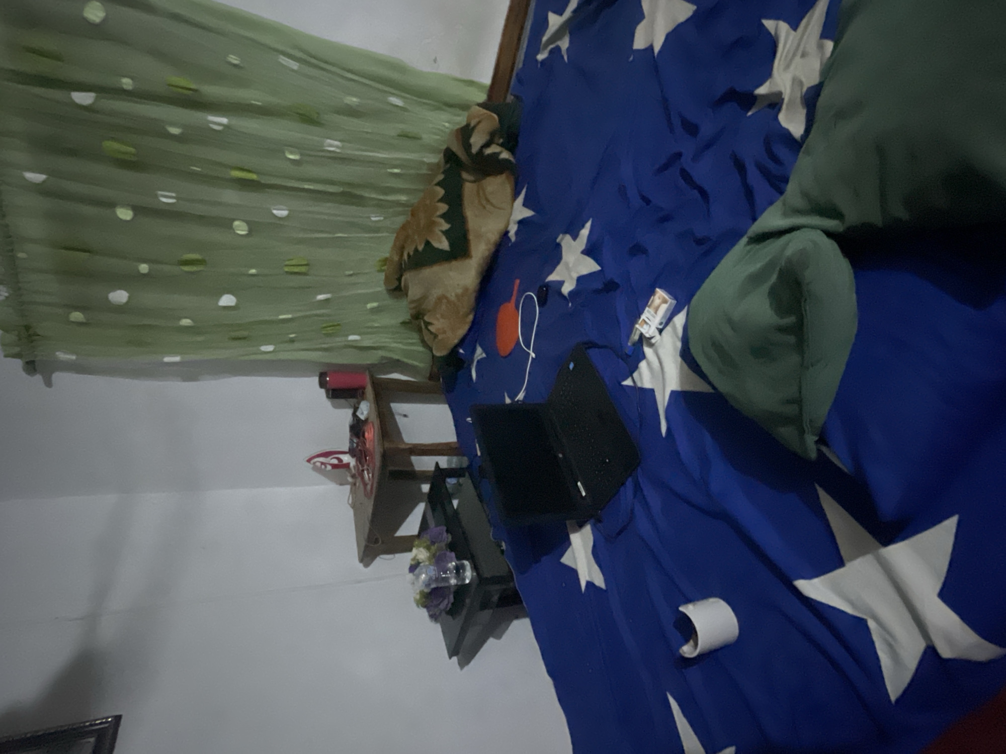 The image depicts a room in Jos, Nigeria. A bed is covered with a blue blanket adorned with white stars, and a green pillow rests on it. A laptop, a pair of orange headphones, and a pack of cigarettes lie on the bedspread. To the side of the bed, a wooden table supports a clock, a potted plant with purple flowers, and a small bottle. A green curtain hangs in the background, and the room's walls are painted white.