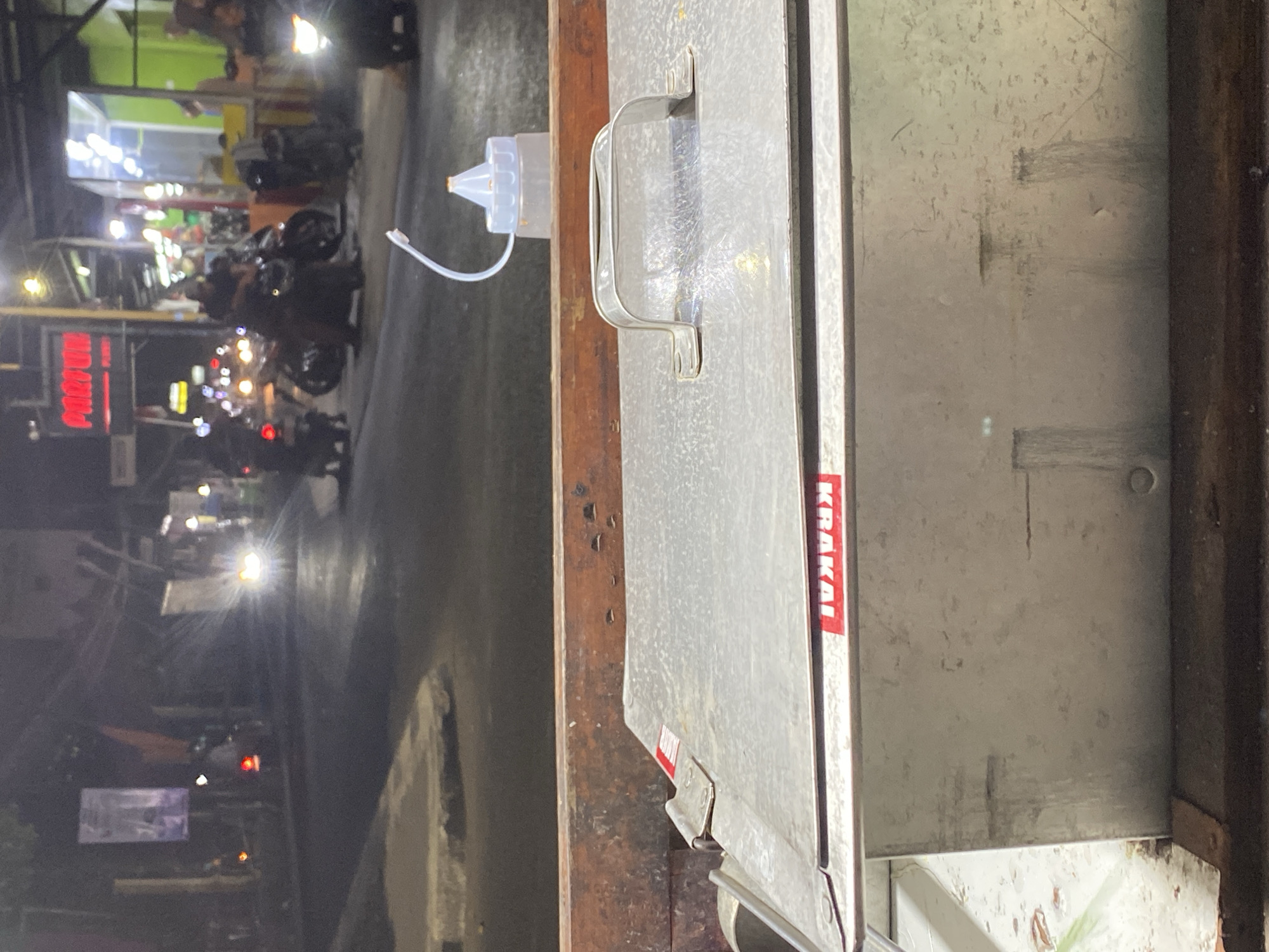 The image depicts a night scene on a street in Kasihan, Indonesia. In the foreground, a metal food cart is visible, its lid closed and displaying a red and white "KRAKAI" sticker. A handle is attached to the lid, and a clear plastic squeeze bottle with a white cap and a clear tube is positioned behind it. The background shows a street with various vehicles, including motorcycles, with their headlights and taillights illuminated. Several buildings with glowing signs and lights are present along the street. The overall impression is a busy urban environment at night, likely a commercial area with street vendors.