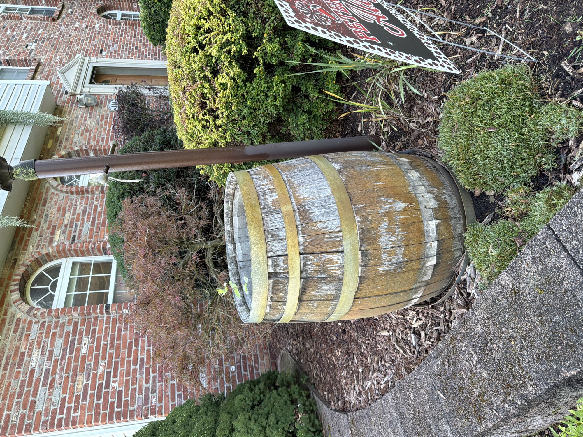 This image depicts an outdoor scene in Peters Township, United States of America, featuring a weathered wooden barrel used as a planter. The barrel is positioned on a bed of dark mulch, adjacent to a stone planter wall on the left. Behind the barrel, a red brick building with an arched window is visible, along with various green shrubs and a Japanese maple tree with reddish-brown foliage. A dark brown, weathered metal pole stands to the right of the barrel. In the foreground, to the right of the barrel, a portion of a sign with red lettering and a black and white spotted border is visible. The overall environment suggests a suburban residential or garden setting.