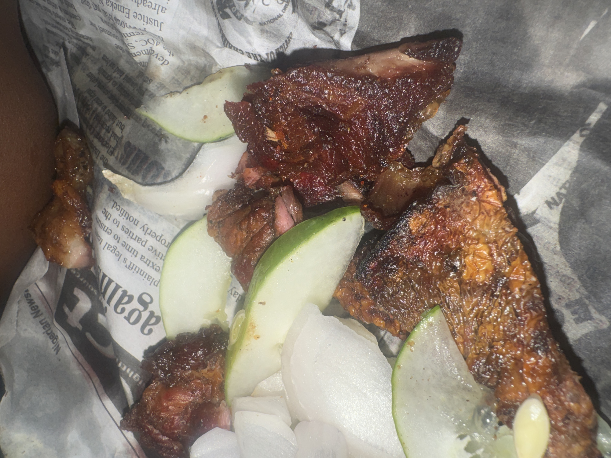 The image shows a close-up, overhead view of a plate of food presented on crumpled newspaper. The food consists of several pieces of what appears to be grilled or fried meat, appearing dark brown and slightly greasy, with some visible seasoning. Interspersed with the meat are slices of pale green fruit, likely cucumber or apple, and chunks of white vegetable, possibly onion or radish. The newspaper beneath the food is printed with text and images, and some of the headlines are partially visible, including "Nigerian News" and "again". The scene is lit from above, creating some highlights and shadows on the food and newspaper. The image does not provide any specific details or context that would allow for the determination of a location within the city of Abuja, Nigeria.