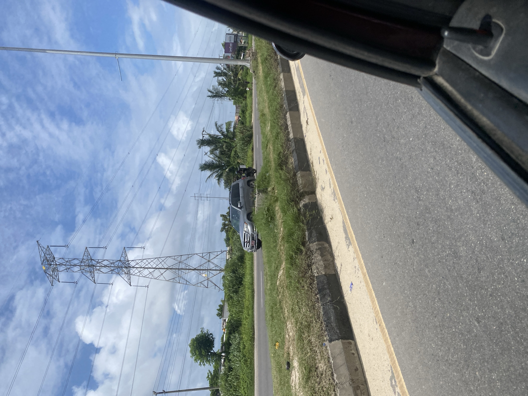 The image shows a view from inside a vehicle, looking out at a road in Lekki Peninsula II, Nigeria. A silver SUV is driving on the road, with lush green vegetation and palm trees lining the sides. A large electricity pylon stands tall in the background, with power lines stretching across the sky. The sky is partly cloudy, with a mix of blue and white clouds. A concrete barrier separates the road from the grassy median. The overall scene depicts a typical roadside view in a tropical urban or suburban environment.
