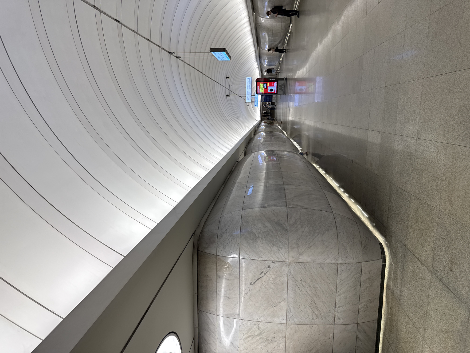 This image captures the modern, brightly lit platform of a subway station. The station features a high, vaulted ceiling constructed from a series of parallel, concave white panels that create a ribbed, arched effect and are uniformly illuminated, providing diffused overhead light. A horizontal, rectangular architectural beam runs along the top of the left wall, separating it from the vaulted ceiling.
The platform walls on both sides are distinctive, appearing as elongated, convex structures clad in polished, light grey marble tiles with natural veining. These curved surfaces are reflective, showing subtle distortions of light and reflections from the opposite side of the platform. A continuous strip of light emanates from the base of these curved walls, illuminating the space between the wall and the floor tiles and emphasizing their sculptural form.
The floor is laid with light grey square tiles that possess a high sheen, reflecting the overhead lighting and the station's architectural elements. In the mid-ground of the platform, a prominent vertical digital display screen with a red background showcases an advertisement featuring Russian text, including "24 999₽," and a graphic of an electronic device. Further down the platform, a smaller, blue-lit digital information screen is suspended from the ceiling.
Several individuals are present on the platform. A small group of people are gathered on the left side, near the large digital screen, while on the right side, two individuals are spaced out, with one appearing to be focused on a mobile phone. The station appears clean and spacious.
This architectural design, characterized by its undulating marble walls and ribbed, illuminated ceiling, is consistent with the **Novoperedelkino station (Новопеределкино)** on the Solntsevskaya line (Line 8A) of the Moscow Metro in Moscow, Russia.