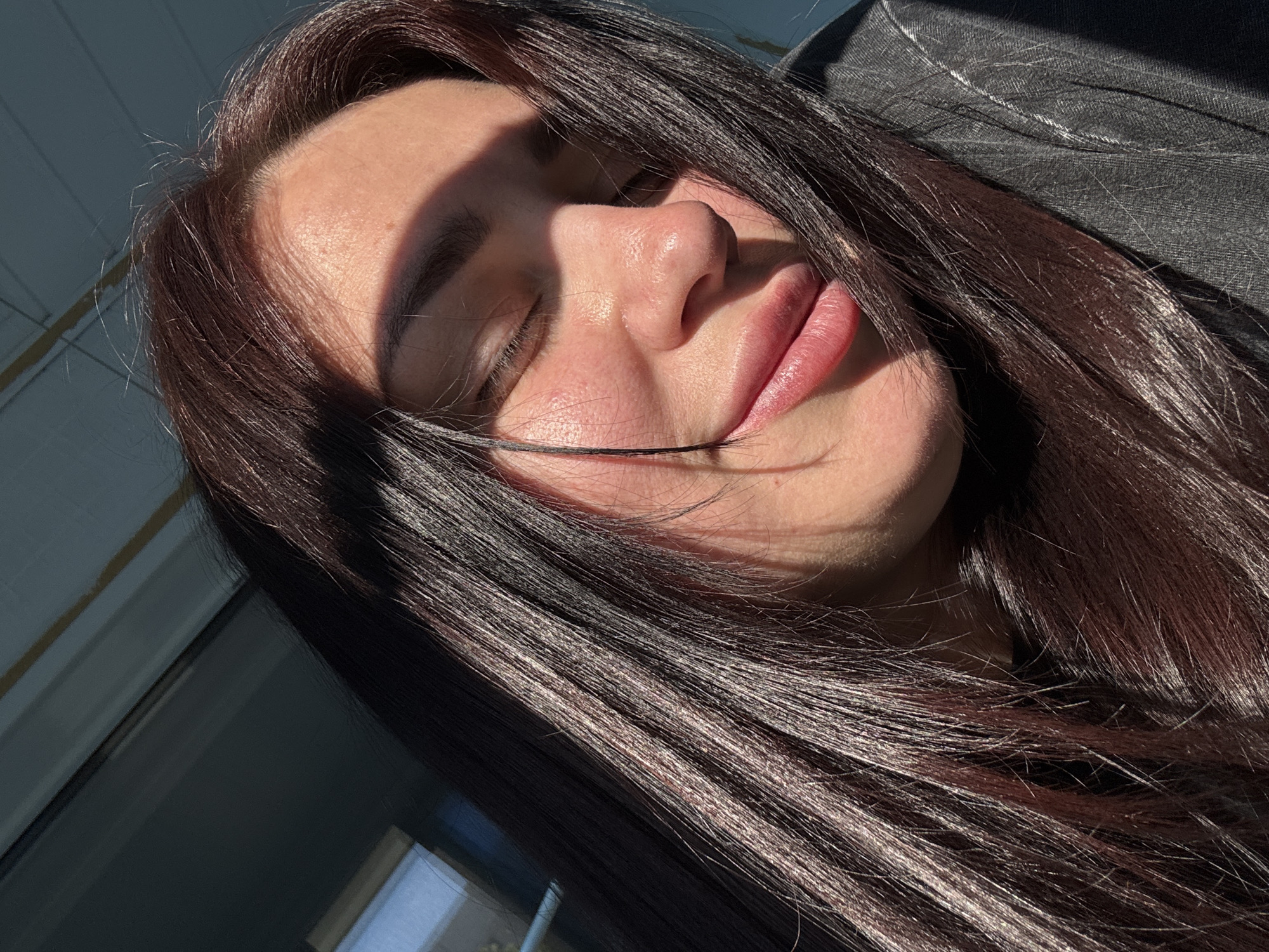 The image shows a close-up of a young woman's face, tilted upwards and to the side. She has dark, long hair that cascades around her face, with some strands falling across her cheek and eye. Her eyes are closed, and her lips are slightly parted, suggesting a relaxed or contemplative mood. Sunlight illuminates her face, casting shadows that highlight her features, particularly her cheekbones and nose. The lighting suggests it is daytime, possibly late afternoon due to the warm glow and long shadows.
She is wearing a dark, textured garment, possibly denim or a similar fabric, with visible stitching details, likely a jacket or shirt. The background is out of focus but appears to be an indoor setting with a window or reflective surface, indicated by muted blue and white tones and faint lines. The overall impression is one of a candid, personal moment captured in natural light. There is no visible text in the image.