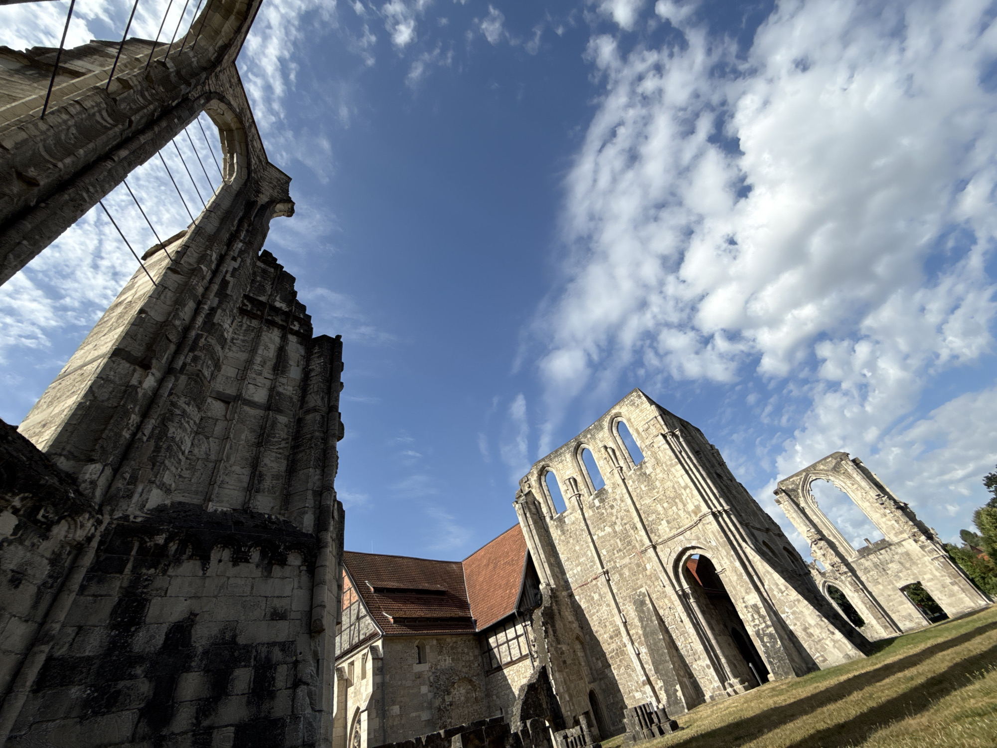 An exterior, low-angle, wide-shot shows the ruins of a stone monastery under a bright blue sky with scattered white clouds. The ruins are composed of large, weathered stone blocks. On the left, a tall, imposing stone structure with large arched openings stands diagonally across the frame. In the center, a section of the monastery with a red-tiled roof and timbered walls is visible. To the right, another section of stone ruins with arched window frames is present. The ground in the foreground is covered with dry grass and slopes upwards. The scene appears to be an outdoor historical site, possibly in the city of Walkenried, Germany, given its architectural style and the presence of historical ruins. There are no people or animals visible in the image.