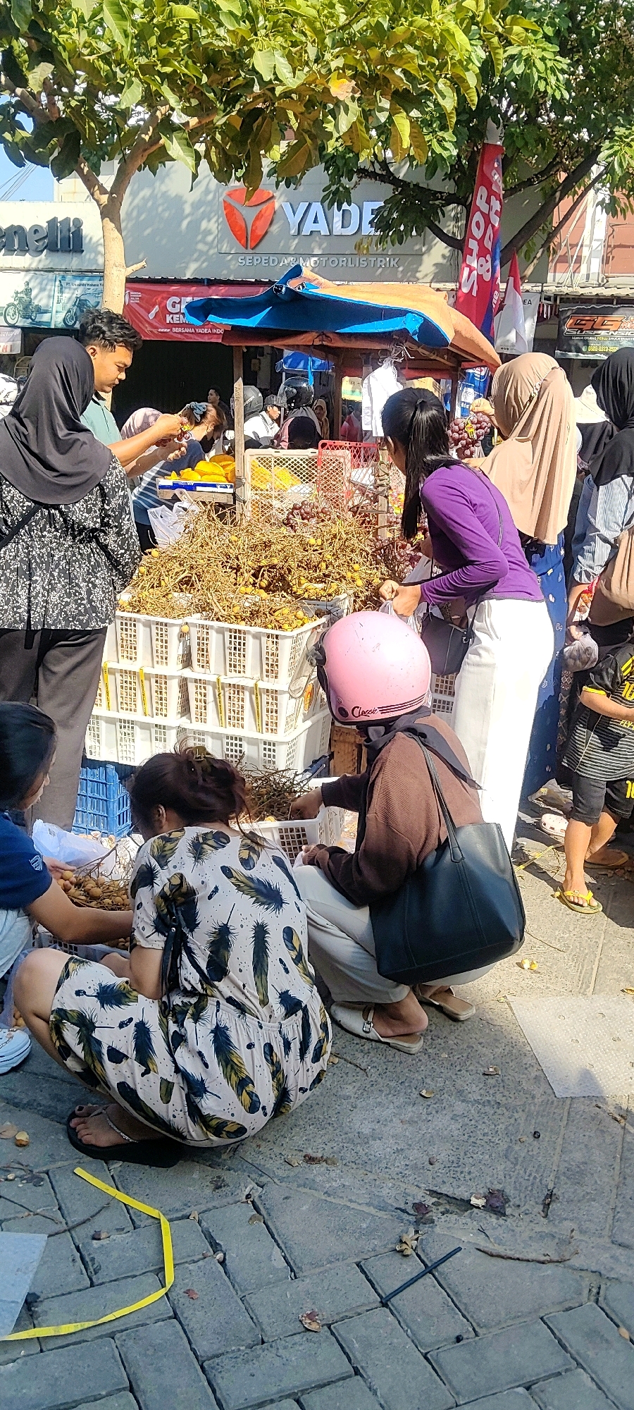 In Cibatu, Indonesia, a street market scene unfolds under the shade of a leafy tree, with a visible "YADEA SEPEDA MOTORLISTRIK" sign indicating an electric motorcycle dealership. The foreground features a selection of fresh produce, primarily grapes and what appear to be longans, displayed in stacked white plastic crates. Several individuals are engaged in the market activities. A woman in a black hijab and patterned blouse stands to the left, interacting with a man in a teal shirt. Nearby, a woman in a purple top and white pants is examining produce, while another woman, partially obscured, is also looking at the offerings. A child in a striped shirt and shorts is visible on the right, standing near a market stall. The ground is paved with grey interlocking bricks, with fallen leaves and small debris scattered about. A pink helmet rests on a surface in the mid-ground. The overall atmosphere suggests a busy, everyday market day.
