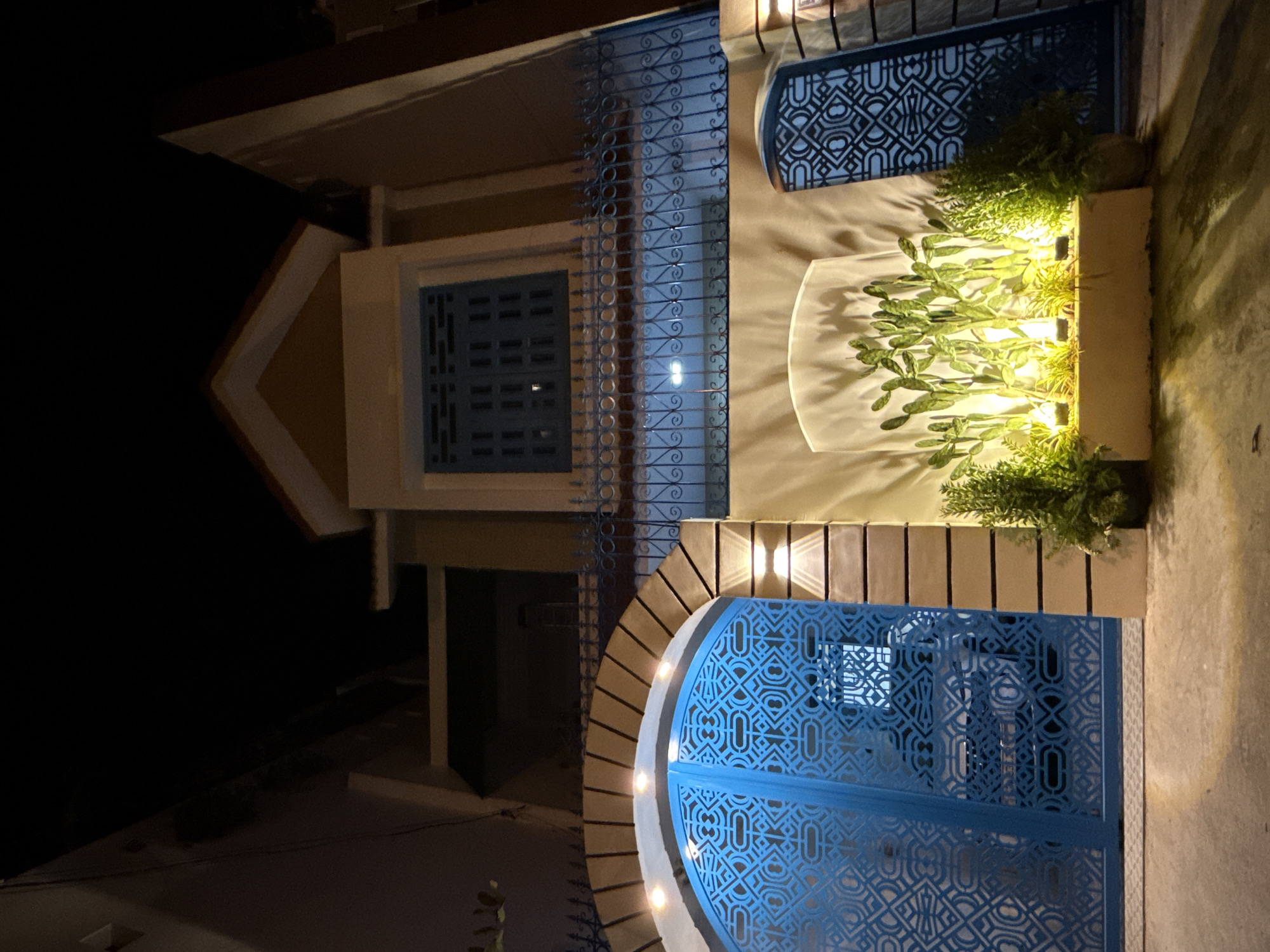 This media file shows an exterior view of a building at night. The building features a prominent blue metal gate with an intricate geometric pattern and decorative blue metal bars on windows. There are also two small outdoor wall lights that cast a warm glow on the facade. In front of the building, a planter box contains several green plants, illuminated from below. The lighting suggests it is nighttime. The scene appears to be a residential or commercial building, possibly a house or a shop, with decorative architectural elements. The overall impression is one of evening illumination and decorative design.