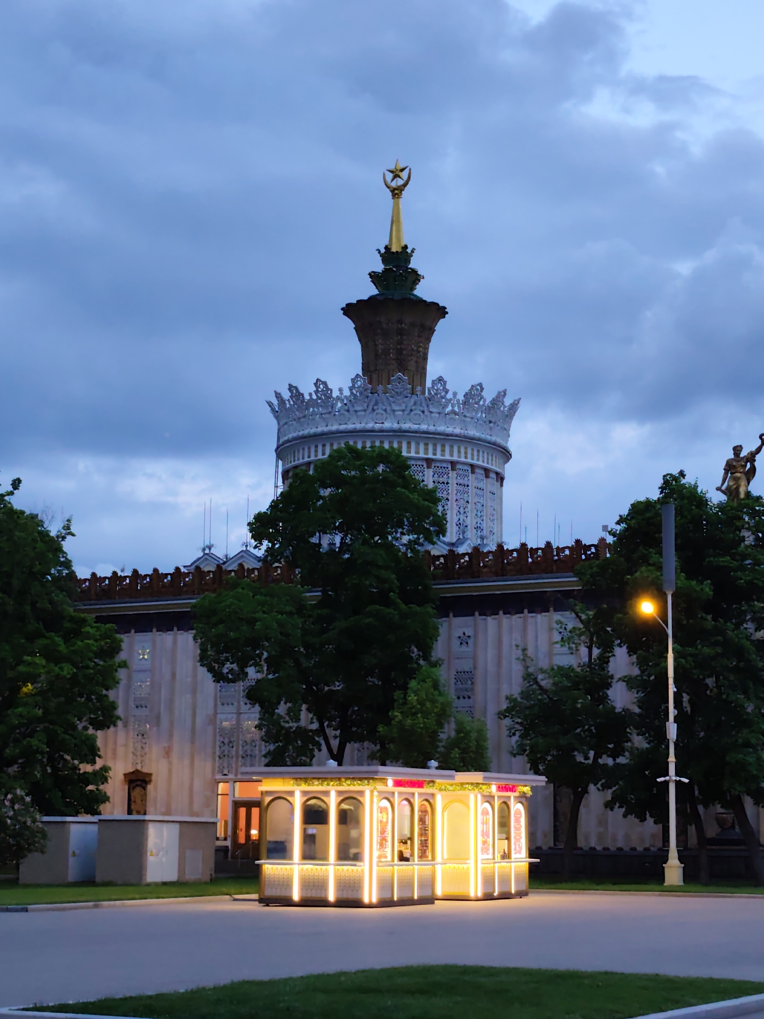 The image depicts a night scene in Moscow, Russia. In the foreground, a brightly lit kiosk with arched openings stands on a paved area surrounded by grass. Behind the kiosk are trees partially obscuring a large building. The building has a tower topped with a golden star and other decorative elements, including a crown-like structure. To the right of the tower, atop the building, is a sculpture of a man holding an object aloft. A streetlight is also visible to the right. The sky is cloudy, suggesting it is dusk or night.