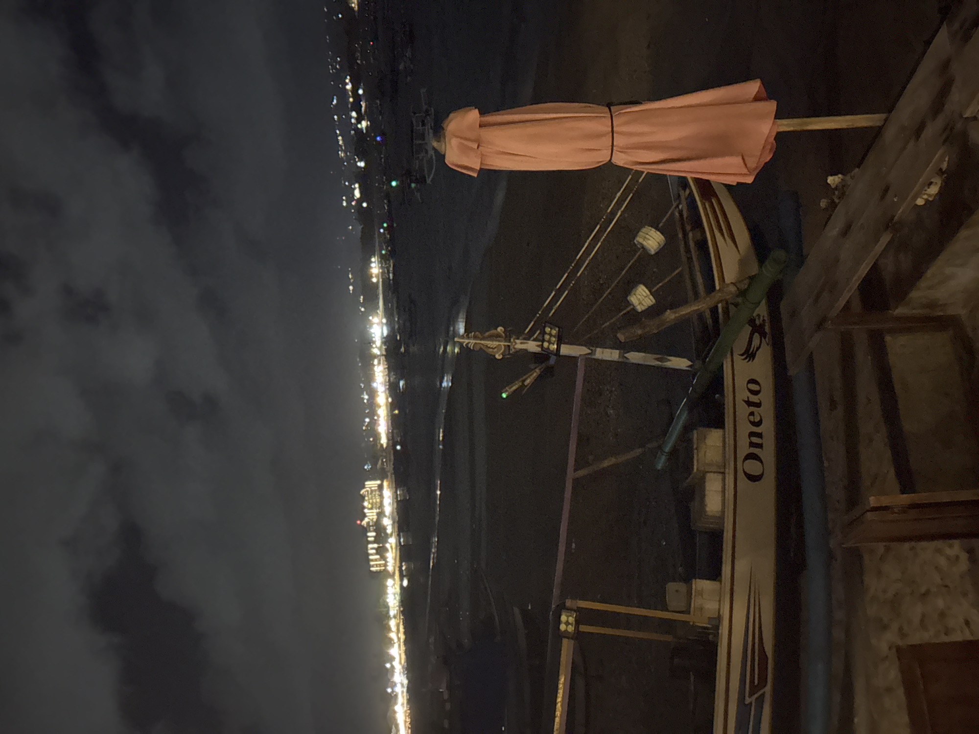 At night in Kuta, Indonesia, a narrow motorized outrigger boat named "Oneto" rests near a beachside structure. The boat features a light pink umbrella and is outfitted with outriggers and rigging. The city lights stretch along the horizon, suggesting a coastal urban environment. The sky is overcast.