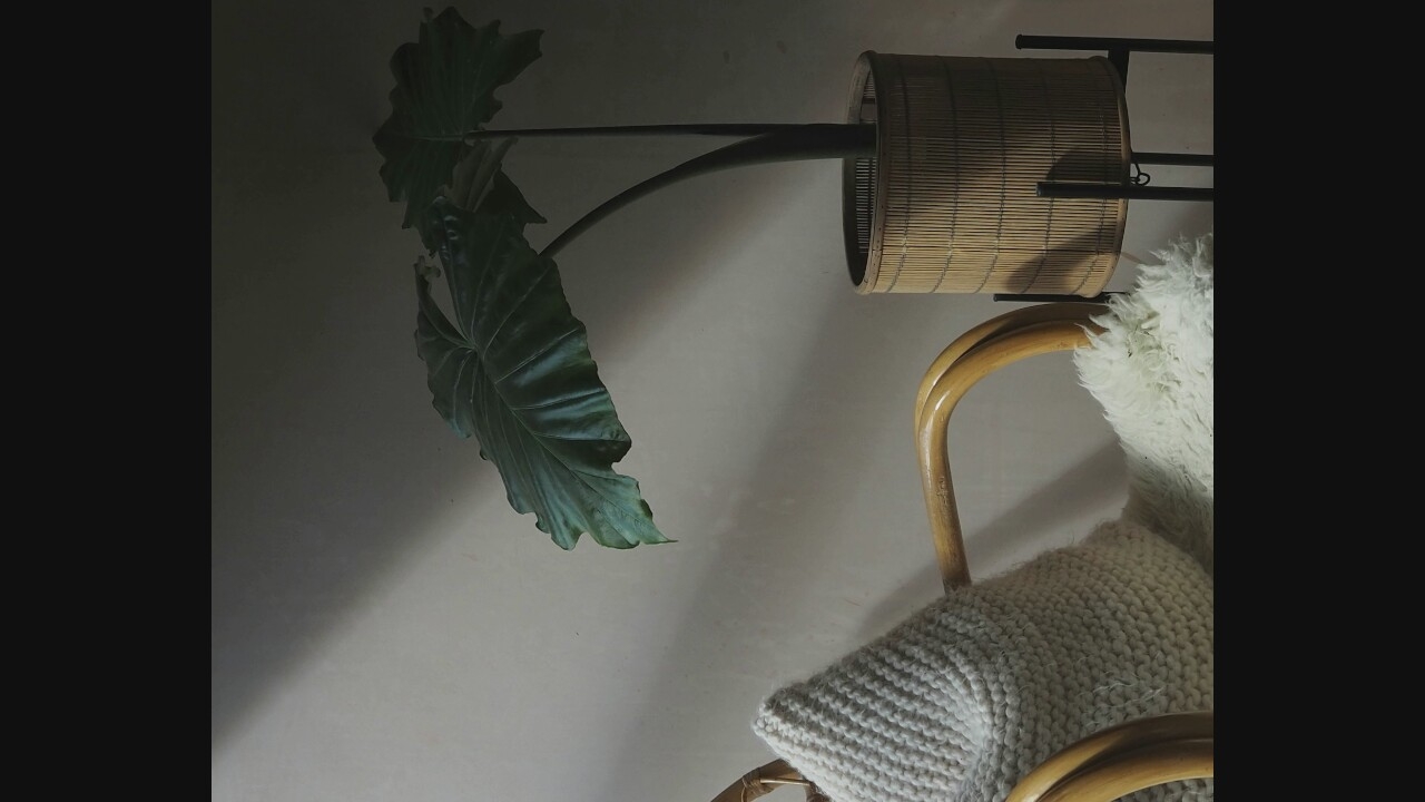 In an indoor setting with a neutral-toned wall, a large houseplant with broad, dark green leaves is situated in a woven cylindrical planter. The planter rests on a dark metal stand. To the left of the plant, a part of a rattan chair is visible, adorned with a large, textured, light-colored cushion and a fluffy white throw. The scene is lit with dramatic shadows cast by an unseen light source, creating a moody ambiance. The location is listed as Unknown, Unknown.