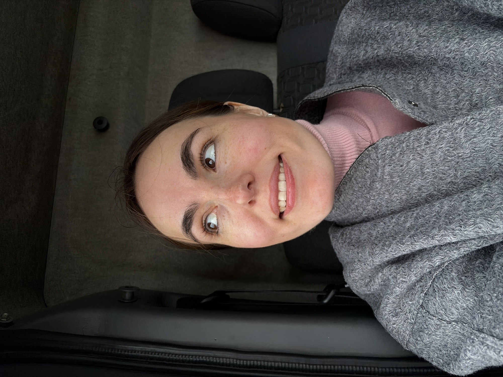 A woman with brown hair and light skin is positioned in what appears to be the trunk of a vehicle. She is wearing a pink turtleneck sweater and a gray wool coat. The interior of the vehicle is gray and black. The vehicle is likely in Izhevsk, Russia, though this cannot be confirmed based on the image.