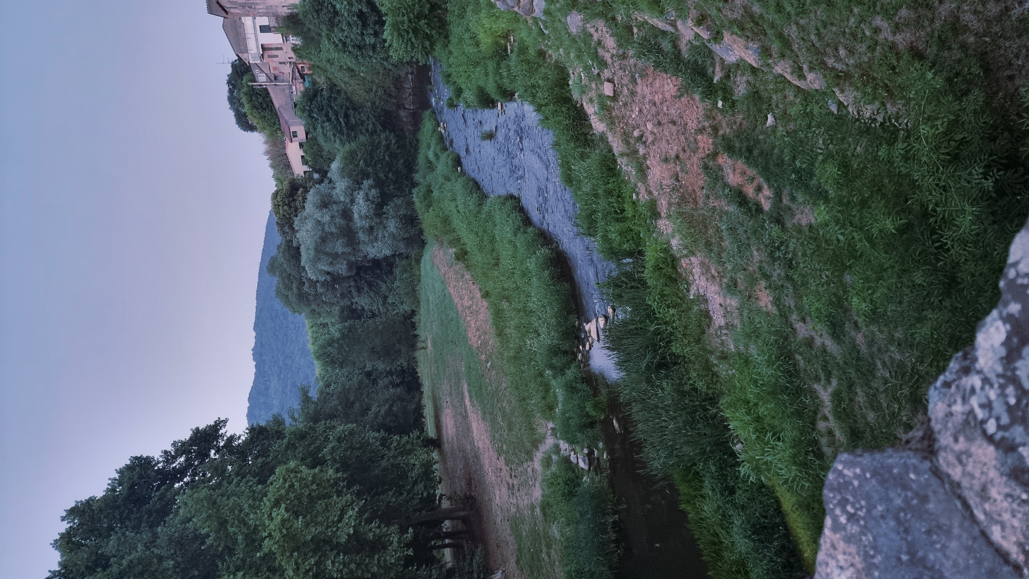A high-angle, long shot captures a winding river flowing through a verdant landscape at dusk. The river, dark and rippled, is flanked by lush green grass and reeds. On the left bank, a grassy clearing is bordered by dense trees. Across the river, on the right bank, the terrain rises to reveal a cluster of buildings, their facades muted in the twilight. Beyond the immediate landscape, rolling hills are visible in the distance, their contours softened by the fading light. The sky above is a pale, dusky blue. The scene is situated in Sant Joan Les Fonts, Spain.