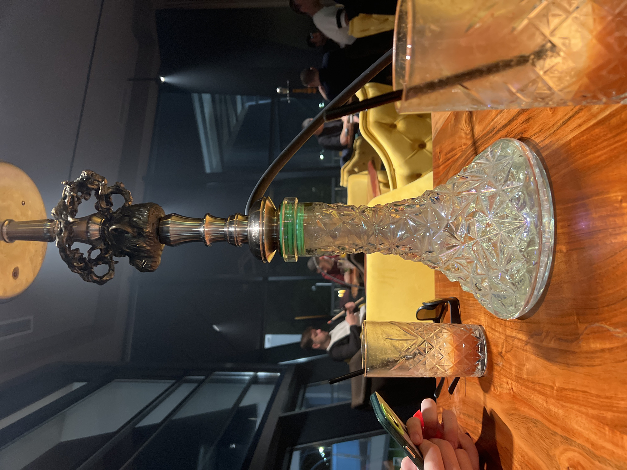 The image depicts a hookah setup on a wooden table in what appears to be a lounge or bar setting. A clear, crystal-patterned hookah base sits on the table next to two patterned glasses, one upright and one on its side. A pair of sunglasses is placed near the base of the hookah. In the background, there are seated patrons, some of whom are smoking. The setting has large windows, and the lighting suggests an evening or indoor ambiance. A phone is also being held by a person whose hands are visible. While the city cannot be definitively determined from the image alone, based on the user's prompt, it can be inferred the location is in Burgas, Bulgaria.