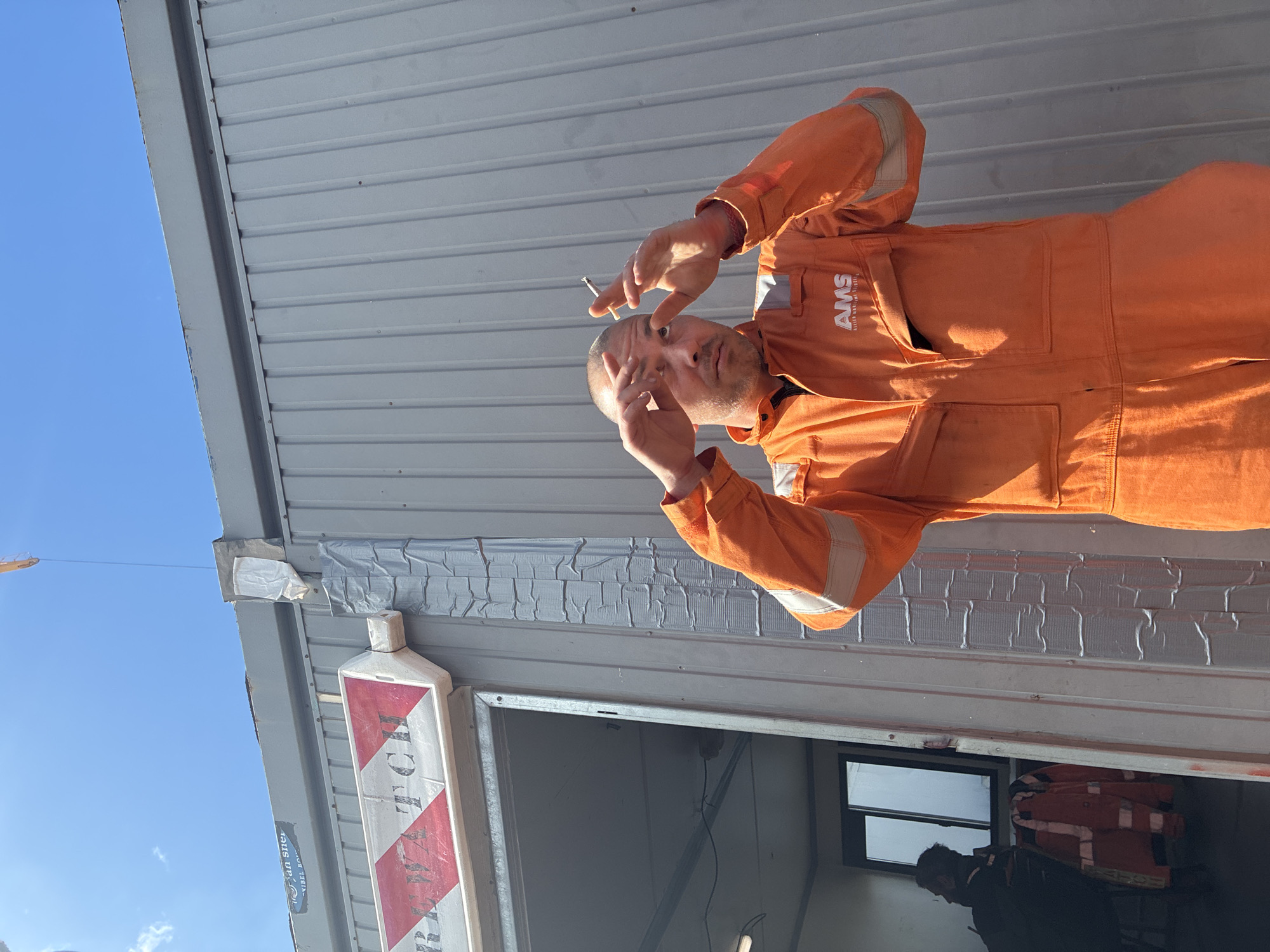 A medium shot, taken outdoors and rotated 90 degrees counter-clockwise, depicts a man smoking a cigarette. He is wearing an orange work jumpsuit with reflective strips on the sleeves. The jumpsuit has the letters "AMS" on the chest. He holds the cigarette between the fingers of his right hand, while his left hand is raised near his forehead. The man is standing in front of a corrugated metal building. Above the door is duct tape. A red and white sign is visible to the left. The sky is blue in the upper left corner of the frame. Inside the building, a person is wearing a dark-colored shirt and standing near a window. Orange jackets are inside. The location is not identifiable from the image, however, I will include “Brest, France” as requested.