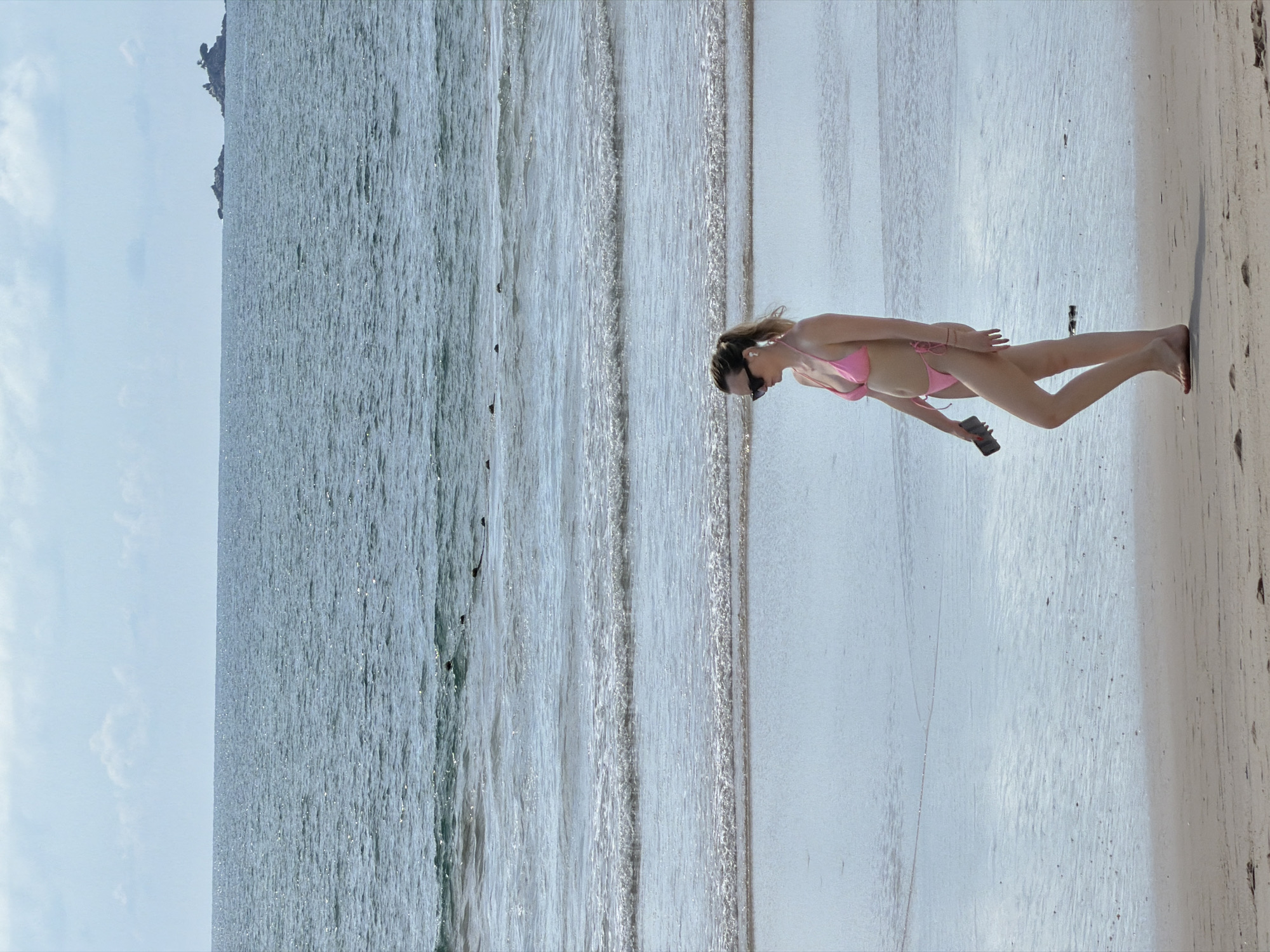 A full-body, medium shot captures a woman from the waist down, walking right to left on a wet sandy beach. She is wearing a pink bikini, sunglasses, and white wireless earbuds. Her right hand holds a dark-colored smartphone. The ocean stretches out behind her, with gentle waves lapping at the shore. The sky above is light blue with scattered white clouds. In the far distance, two small rocky islands are visible on the horizon. The scene appears to be located on a beach in Choeng Thale, Thailand, given the typical tropical coastal environment.