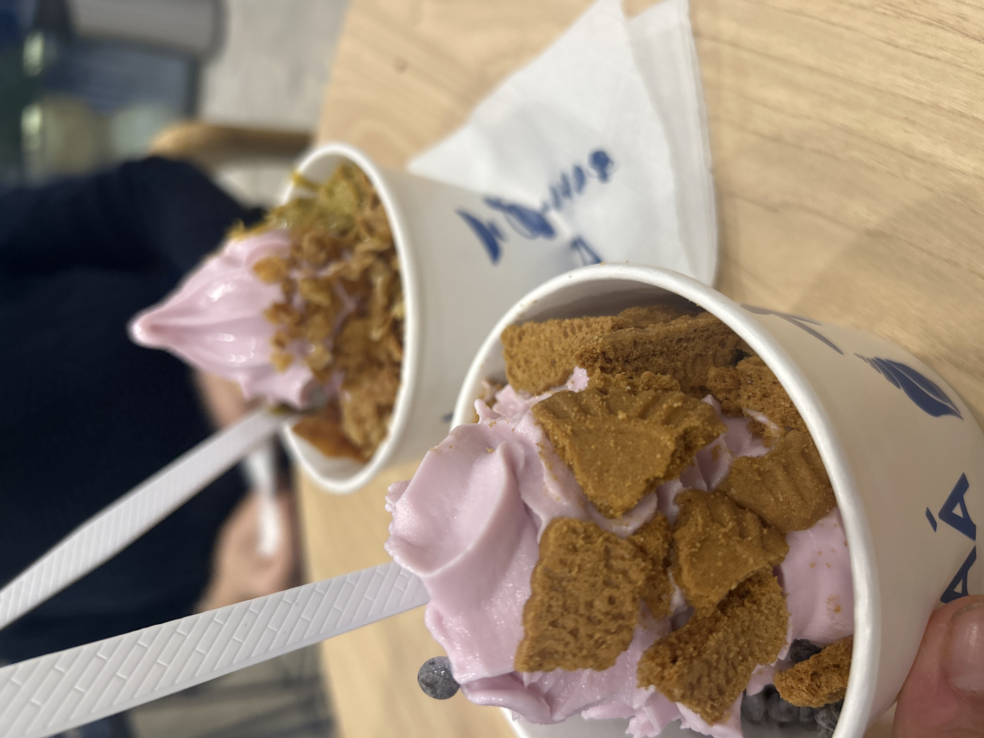 Two cups of frozen yogurt are on a light brown table. The frozen yogurt is light purple and topped with graham cracker-like squares and cereal, and are served with white plastic spoons. A white napkin sits between the cups. The scene is likely indoors, in a food service establishment. There is no verifiable evidence to suggest the location is San Bernardino Tlaxcalancingo, Mexico.