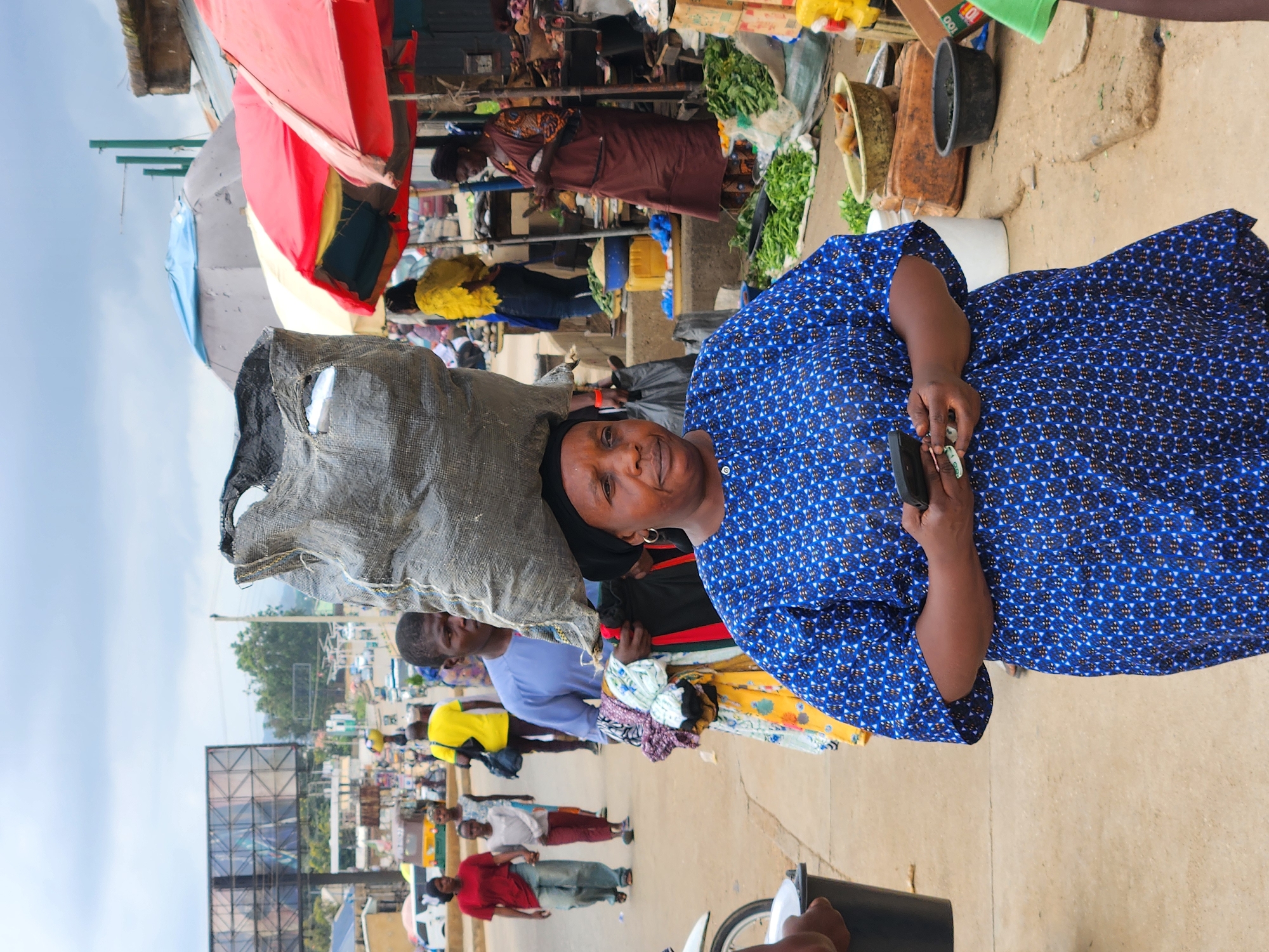 The image captures a bustling outdoor market scene, likely in Kabong, Nigeria, filled with people and commerce. The perspective is at eye-level, focusing on a woman in the foreground.
The central subject is a woman with dark skin, positioned slightly to the right, looking directly at the camera with a gentle smile. She wears a flowing blue dress patterned with white and black geometric shapes. A large, grey, woven sack, possibly containing merchandise or personal belongings, is expertly balanced on her head. Her right hand, raised to her chest, holds a small, dark object, perhaps a mobile phone, along with a small, lighter-colored item between her fingers.
Behind her, the market is vibrant with activity. Numerous individuals are visible, either tending to stalls or moving through the marketplace. Several market stalls are set up, featuring colorful umbrellas in red, blue, yellow, and grey, providing shade. Green leafy vegetables are displayed in baskets at some of these stalls. Piles of fabric or clothing are also seen among the goods. Various containers, including black, yellow, and brown buckets, are scattered around the stalls.
The background reveals more of the market environment, with people dressed in diverse clothing, including yellow and light blue shirts. The ground consists of both dirt and paved surfaces. In the mid-distance on the left, a large, empty billboard structure is prominent, with a street and buildings extending further back. Utility lines are visible against the sky. A portion of a motorcycle or scooter is visible in the lower right.
The scene depicts a lively daytime setting, with a bright, slightly overcast sky. The woman's expression conveys a sense of calm amidst the market's activity. The only discernible text is "100" on a brown box in the background.