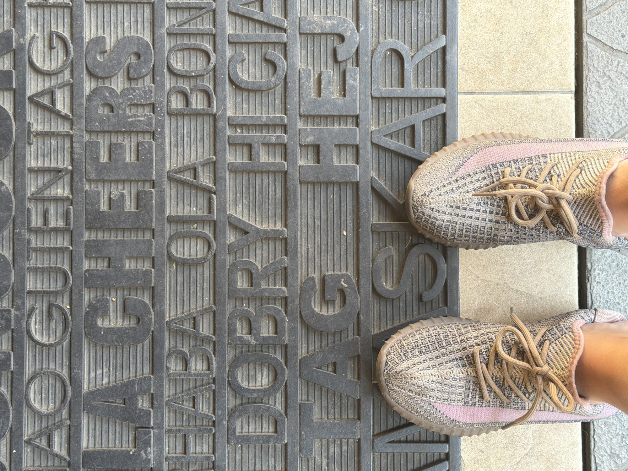 The image is a close-up, overhead shot of a doormat with various greetings in different languages. The doormat is black with embossed text that includes "TAG" and "HEJ". It rests on a tiled surface. In the foreground, two feet wearing patterned sneakers are visible. The doormat appears to be located in the city of Qarağandı, Kazakhstan, likely outside an administration building, as suggested by the presence of greetings in multiple languages.