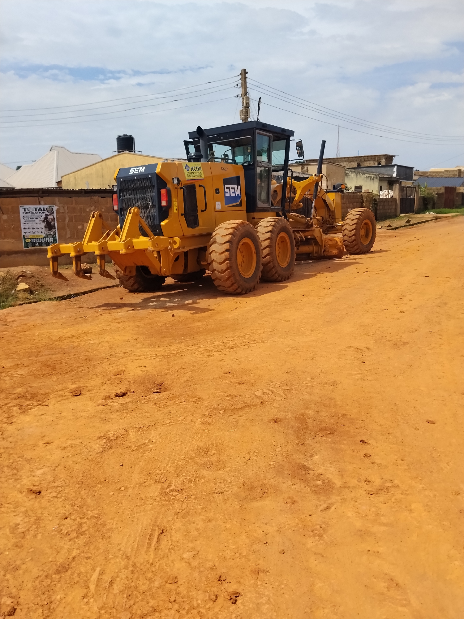 A yellow SEM grader machine is parked on a dirt road in Laminga, Nigeria. The machine is facing towards the right of the image. Behind the grader, there are buildings and a water tank. A poster is attached to a brick wall on the left side of the image. The sky is partly cloudy. The grader has large tires and a blade in the front. The overall scene depicts a construction or road maintenance site.