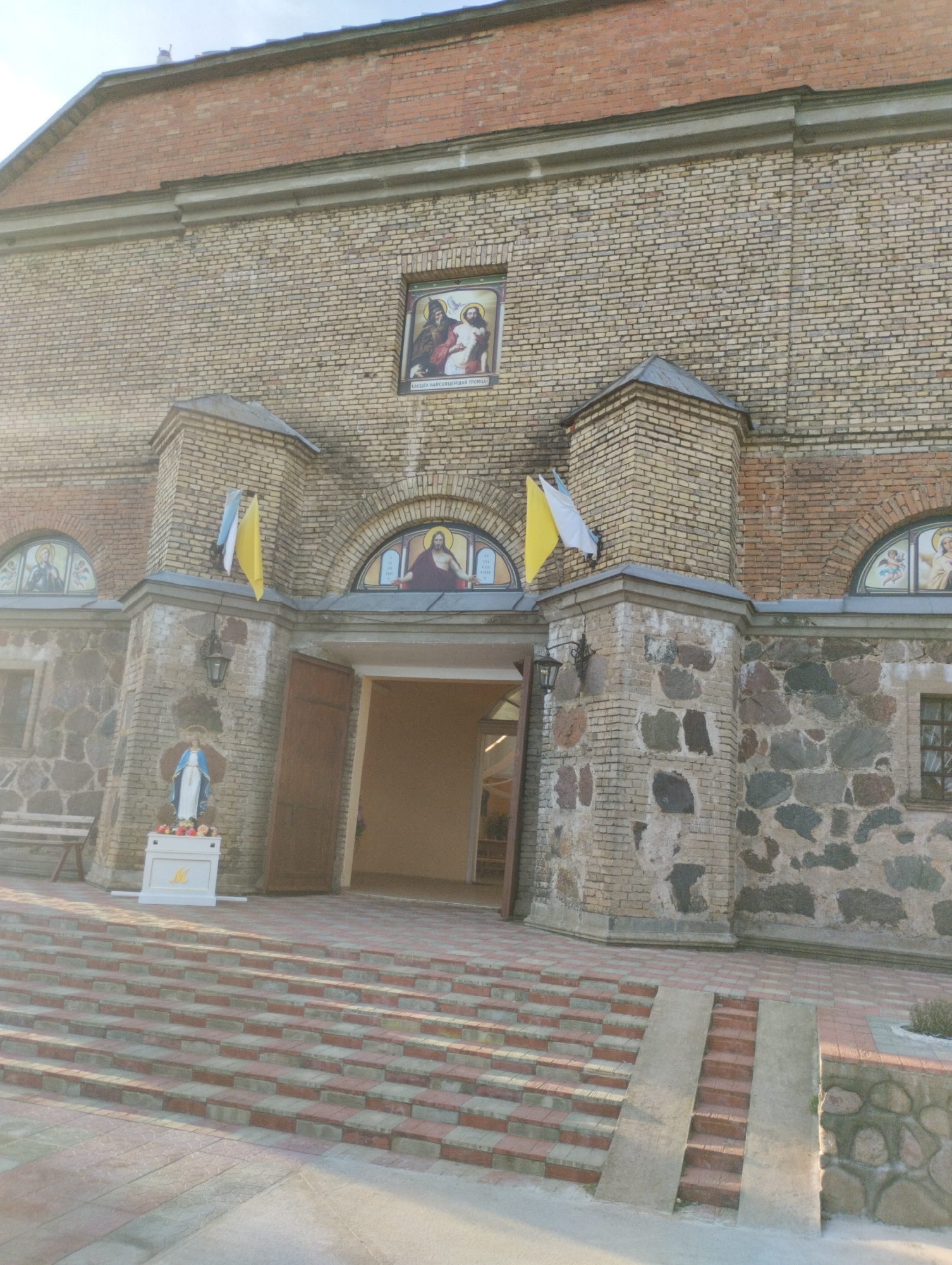 The image shows the exterior of a brick church in Вішнеўскі сельскі Савет, Belarus. The church has a large arched entrance with wooden doors and a set of red and gray brick stairs leading up to it. Above the entrance, there are arched stained-glass windows depicting religious figures. To the left of the entrance, a statue of the Virgin Mary stands on a pedestal. Two flags, one yellow and one white and blue, are hanging from poles on the side of the building. The building's facade is constructed from light-colored bricks and features sections of rough stone masonry. The scene is illuminated by natural light, casting shadows on the stairs and the building.