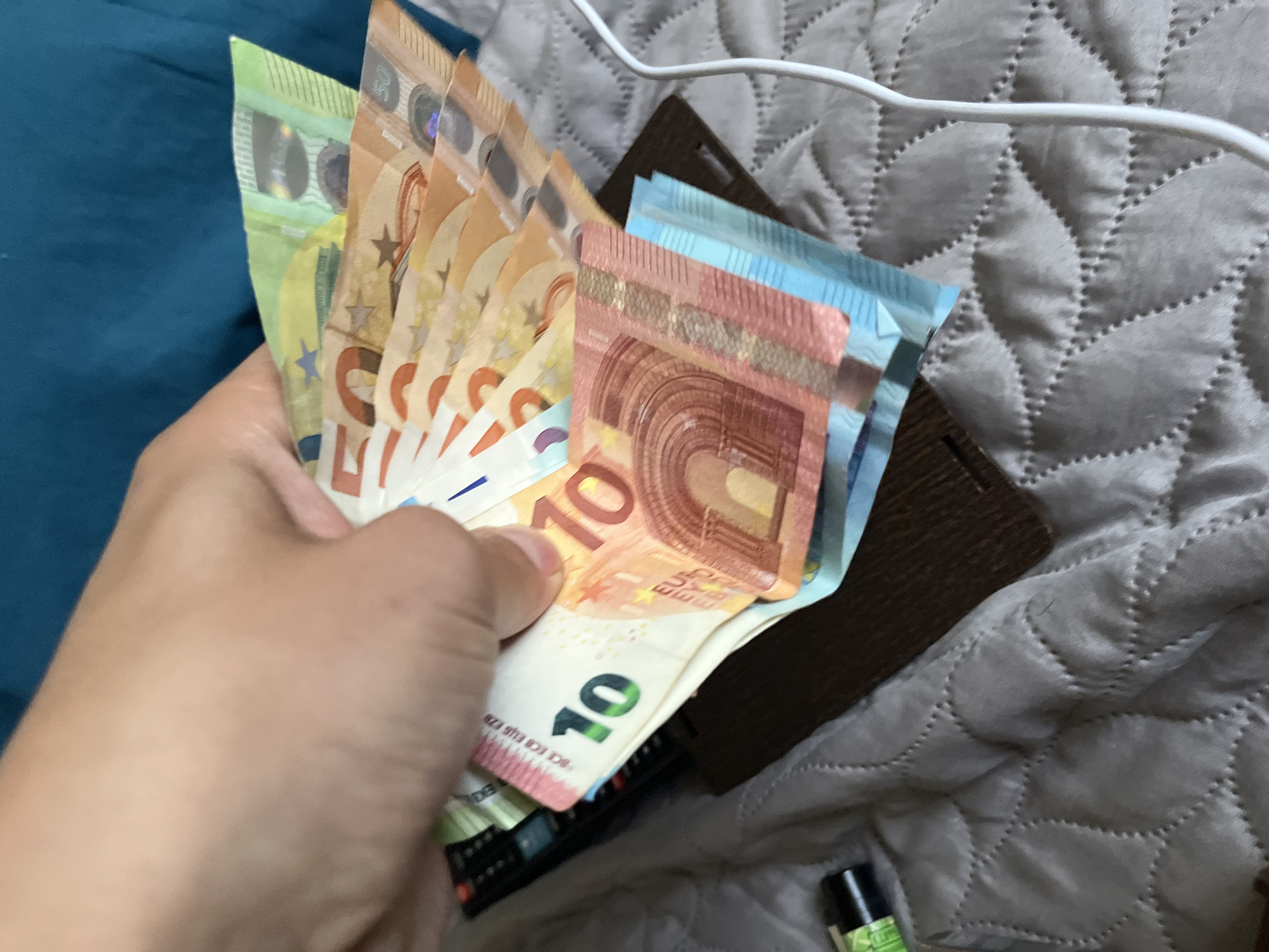 A close-up, eye-level shot captures a person's hand holding a fan of Euro banknotes. The hand, positioned centrally, is displaying various denominations of Euro currency.
Prominently featured in the foreground is a red-brown 10 Euro banknote, clearly showing its "10" denomination and the text "EURO EU EBPO." Behind it, several blue 20 Euro banknotes are partially visible, tucked into a dark brown object that appears to be a wallet or money clip. To the left of the 10 Euro note, the hand holds multiple orange 50 Euro banknotes, with the number "50" discernible on them. Further left, a green 100 Euro banknote is partially seen, indicating a significant sum of money.
The setting appears to be indoors. The hand is positioned over a gray, quilted fabric, likely a blanket or bedspread, characterized by a repeating stitched pattern. A solid teal-colored fabric is visible on the far left. In the upper right of the frame, a white cable runs across the quilted surface. At the bottom left, beneath the hand, a portion of a black remote control with buttons is visible. A small, dark-colored bottle with a green label is also present in the bottom right corner.
The activity shows someone handling money, potentially counting or organizing the banknotes. While Euro currency is depicted, the provided context indicates the location is Sofia, Bulgaria. Visible text includes the denominations "10," "50," "100," and partial bank abbreviations on the Euro notes.