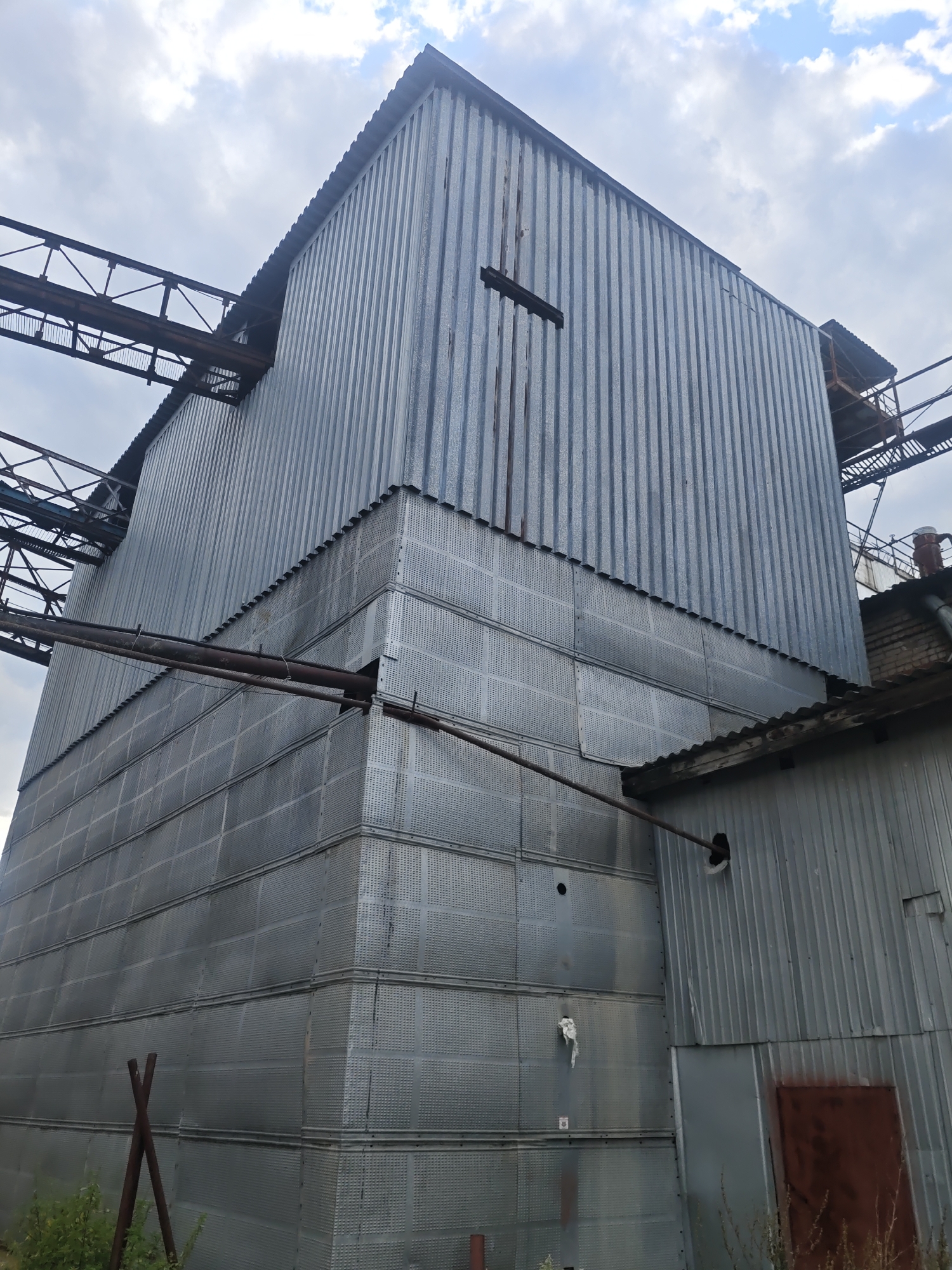 This is a low-angle, full-shot photograph of a large, industrial metal building, likely a granary or silo, located in Platoschino, Russia. The building has a corrugated metal exterior and appears to be quite old and weathered, with signs of rust and some minor damage. A section of the building features a perforated metal cladding, creating a grid-like pattern. Two large, rusty pipes run horizontally across the lower half of the building's facade. Above and to the left of the main structure, a network of metal walkways and supporting beams are visible, extending outwards and upwards, suggesting an interconnected industrial complex. The sky is partly cloudy with a mix of white and gray clouds. In the foreground, some sparse greenery and two vertical metal poles are visible at the base of the building. To the right of the main structure, a smaller, attached structure with a corrugated roof and a dark, rusty metal door is present. The overall scene conveys a sense of industrial decay and abandonment in a rural or semi-industrial setting.