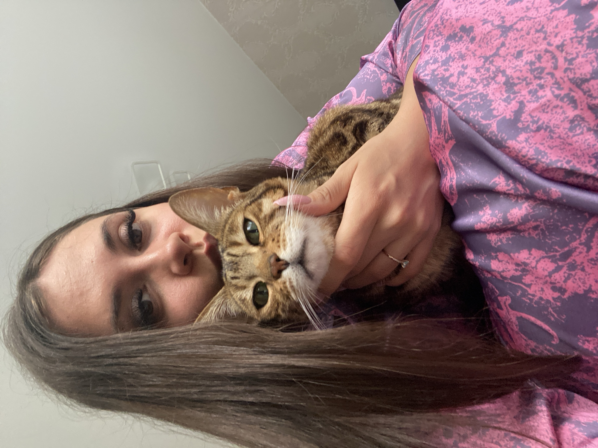 A young adult female is depicted holding a domestic shorthair cat in an indoor setting within the city of Chaikovsky, Russia. The female, with long, straight, dark brown hair, wears noticeable false eyelashes and a light pink/nude manicure. A thin ring with a small, light-colored stone is visible on her left ring finger. She is dressed in a purple garment featuring an abstract pink pattern, possibly a robe or blouse. Her mouth is slightly puckered towards the top of the cat's head, which is partially obscured by her hair. She maintains direct eye contact with the camera. The cat, a tabby-patterned breed with green eyes and prominent white whiskers, is held securely in her arms, also looking directly at the camera. The background consists primarily of a plain light-colored wall and ceiling, with a textured, patterned wall covering partially visible in the upper right quadrant.