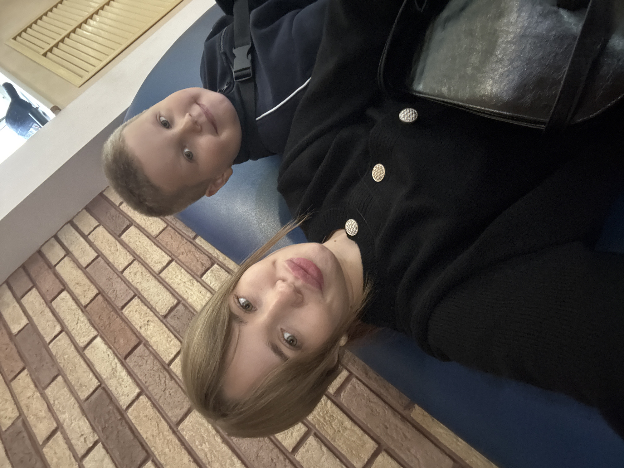 A young woman and a young boy are lying down next to each other on a blue surface. The woman has blonde hair and is wearing a black cardigan with silver buttons. The boy has short brown hair and is wearing a dark blue jacket with a white stripe. A black leather bag is visible next to the woman. The background includes a brick wall and a wooden louvered blind. The image was taken indoors in what appears to be a waiting area or lounge. Based on the user request for location information, it can be inferred that the image was taken in the city of Chaikovsky, Russia.