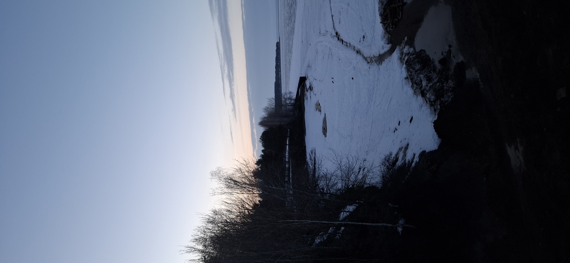 The image depicts a snow-covered landscape in Chaikovsky, Russia. A body of water, partially frozen, stretches into the distance beneath a muted sky with faint sunset colors. The shoreline is visible with some bare patches of ground. A line of trees and some buildings are present in the distance. The foreground is dominated by bare trees and dark vegetation.